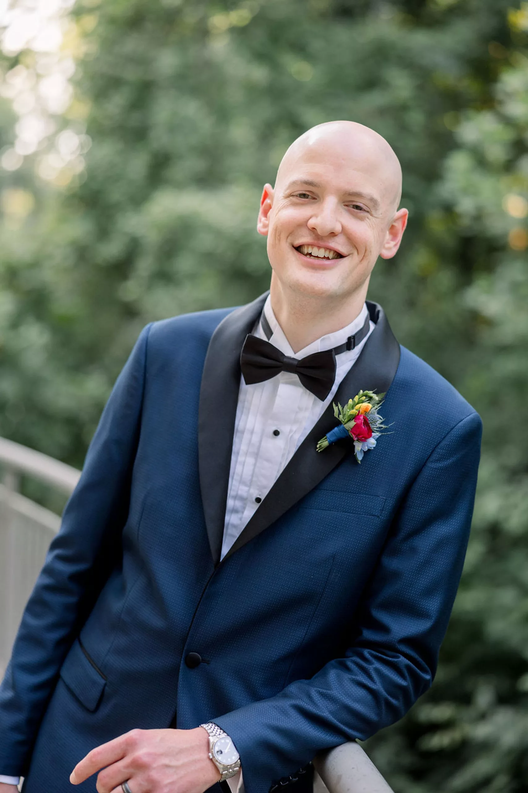 A groom smiles while leaning on a railing outside in a blue tuxedo jacket at his jurassic park wedding