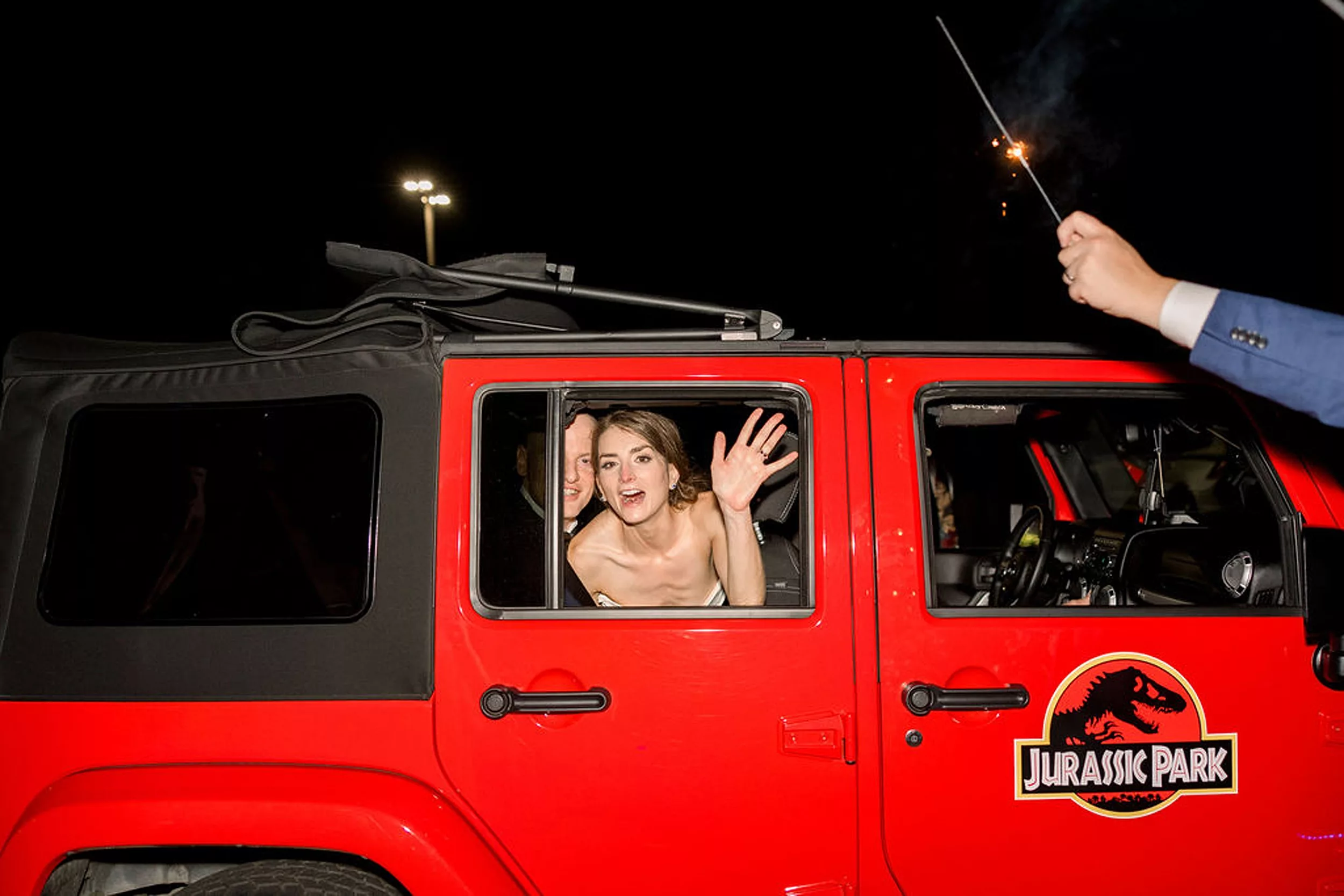 A Red Jurassic Park Jeep leaves a wedding as the bride waves to her guests with sparklers