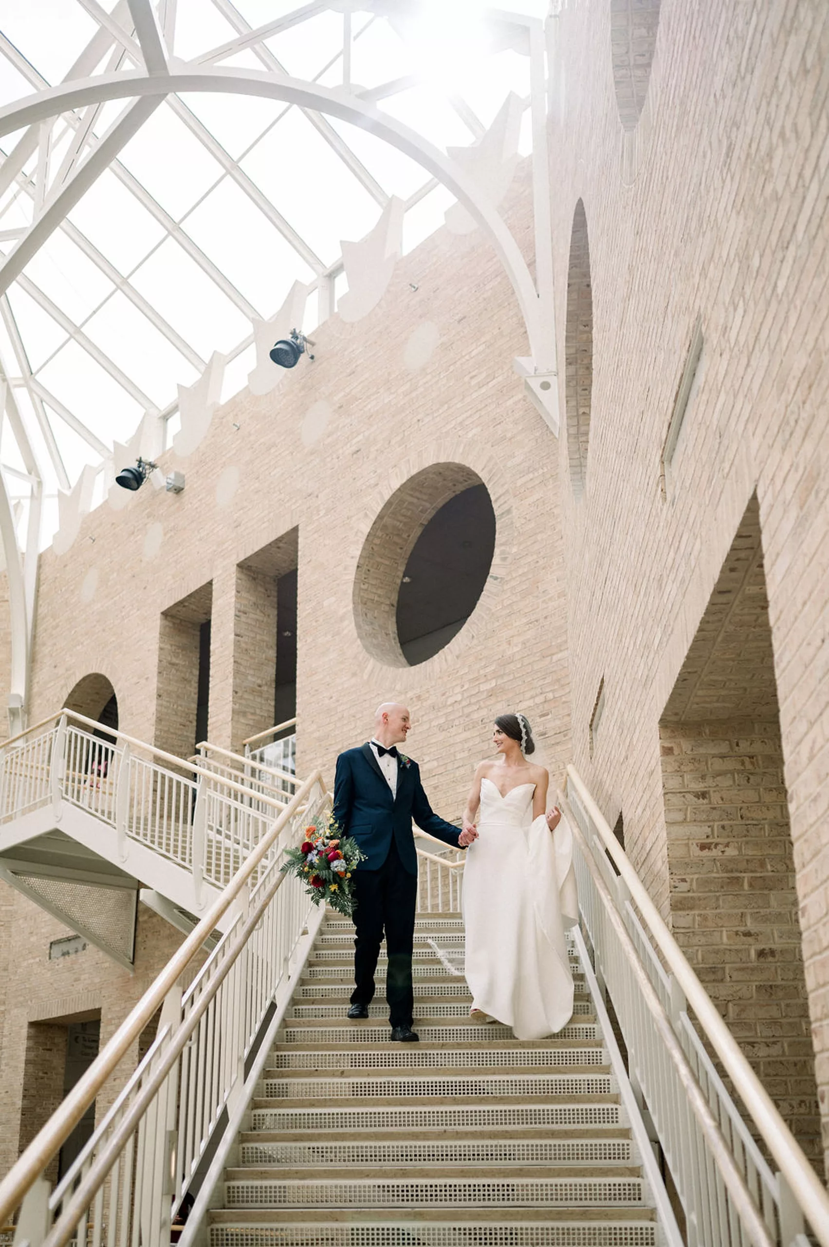 Newlyweds walk down a large museum staircase holding hands