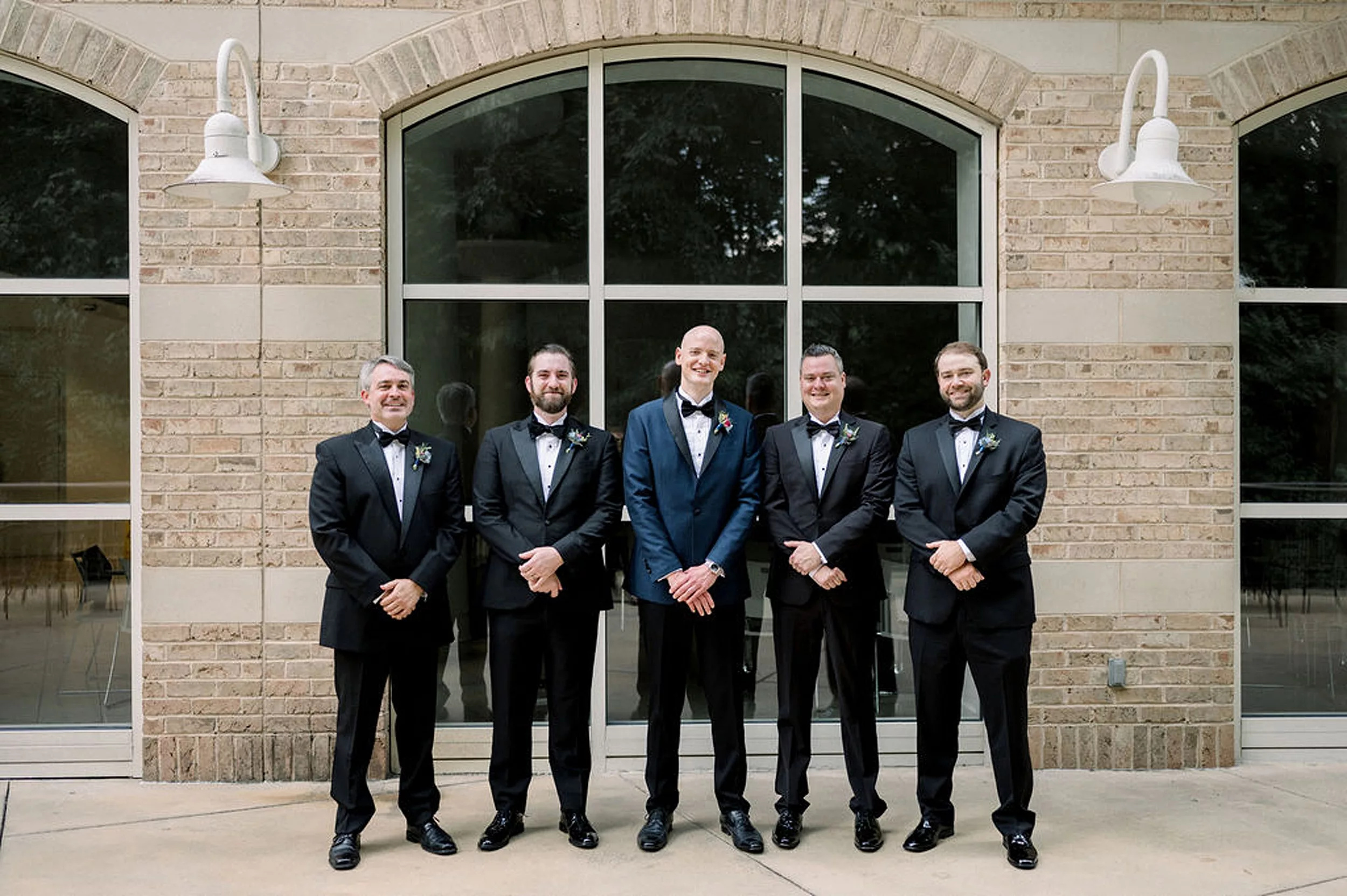 A groom in a blue tuxedo stands with with his groomsmen in black tuxedos in front of large windows of a museum