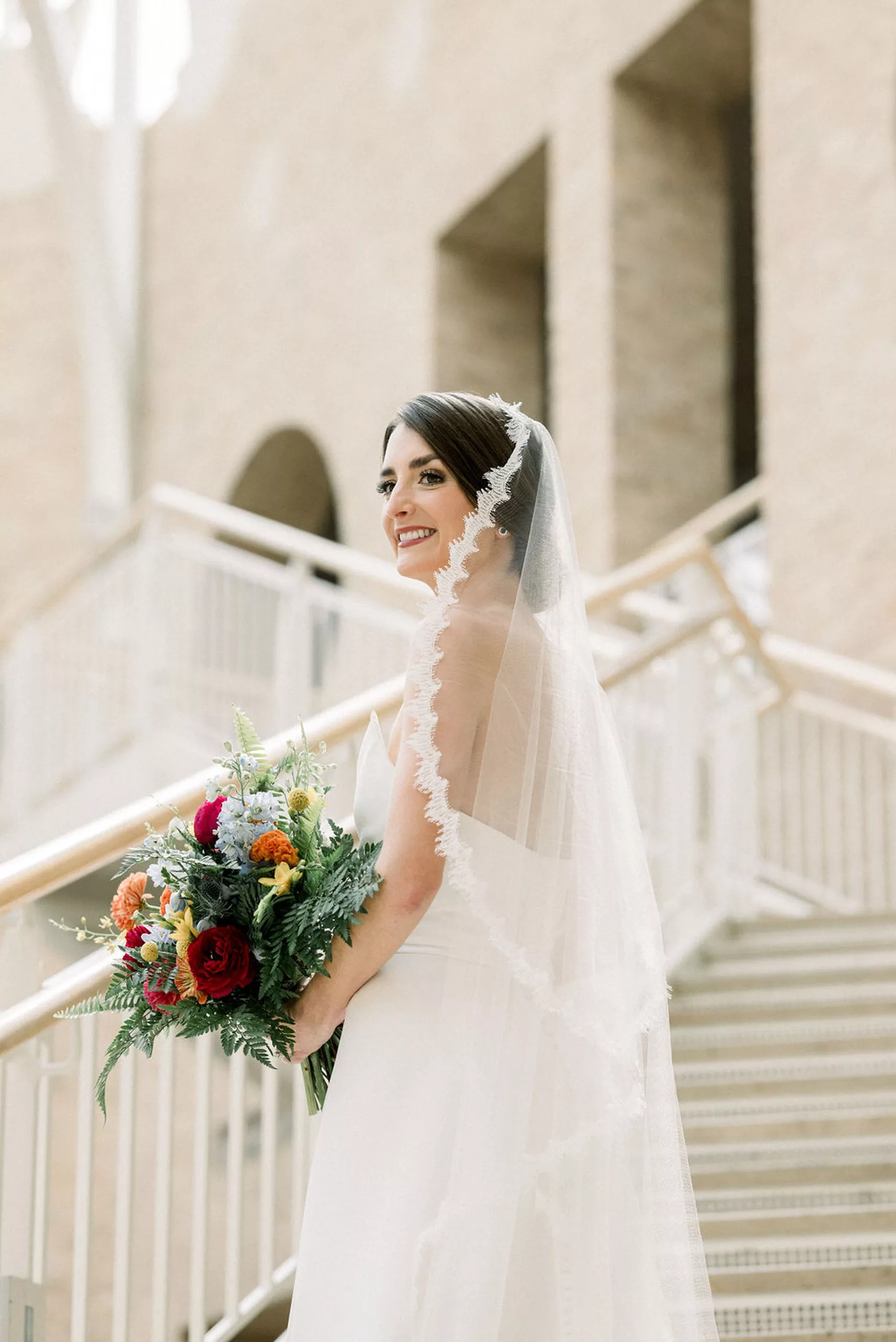A bride walks up a set of stairs smiling over her shoulder in a museum holding her fern and colorful flower bouquet