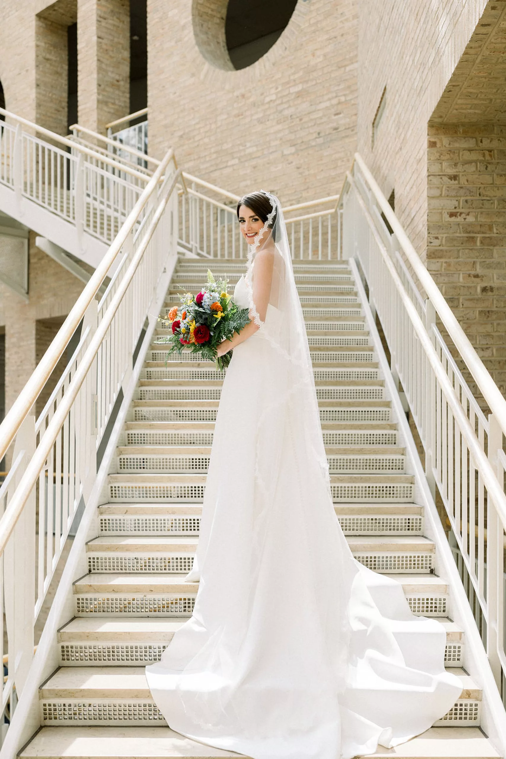 A bride stands in a tall stairway looking over her shoulder with her train flowing behind her holding her colorful bouquet in a museum