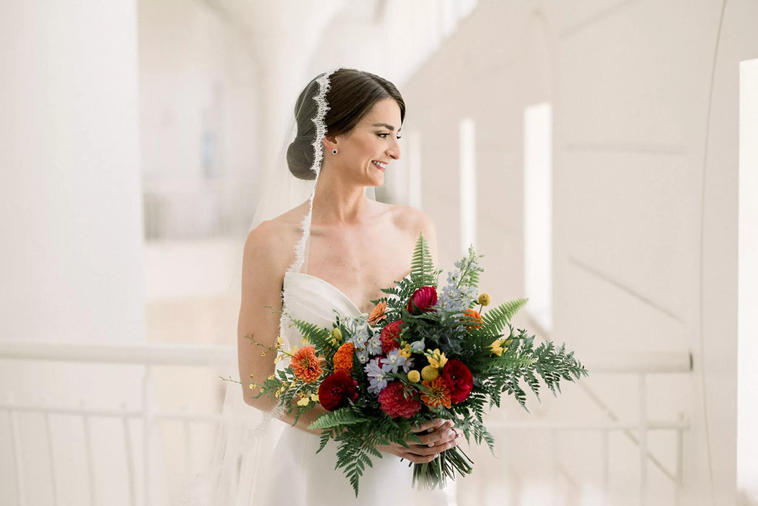 A bride stands looking out a window holding her fern and colorful flower bouquet and lace veil