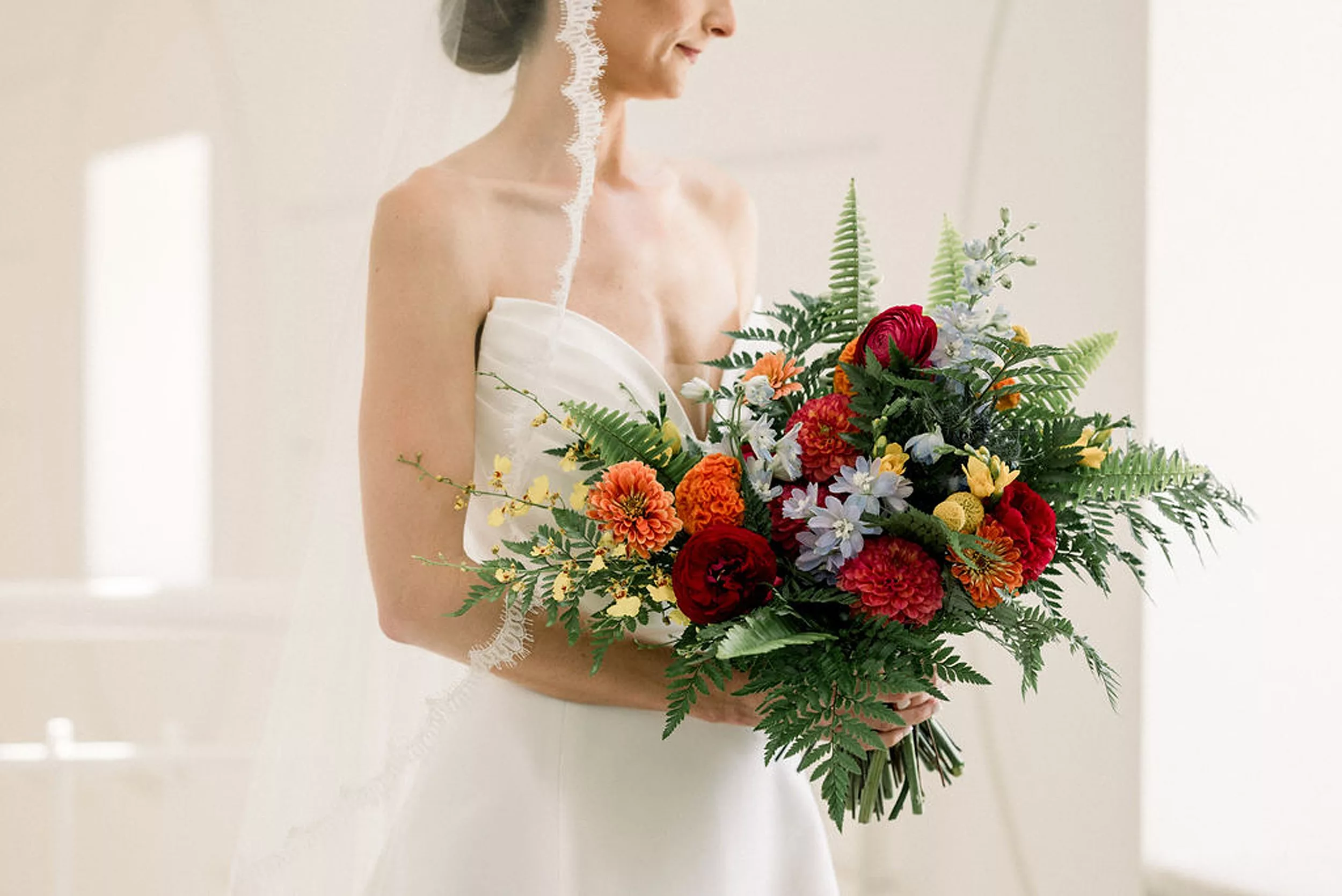 A bride stands in a window in a silk dress holding a bouquet of ferns and colorful flowers