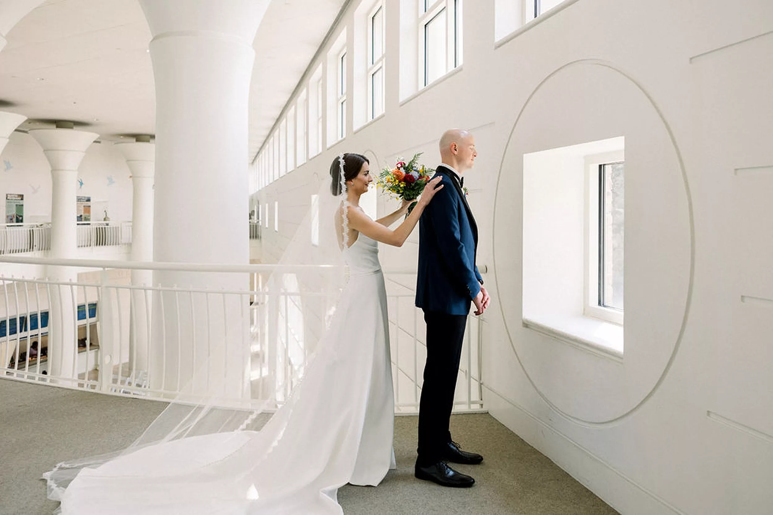 A bride taps the shoulder of her groom for a first look as he looks out a window with his back turned to her