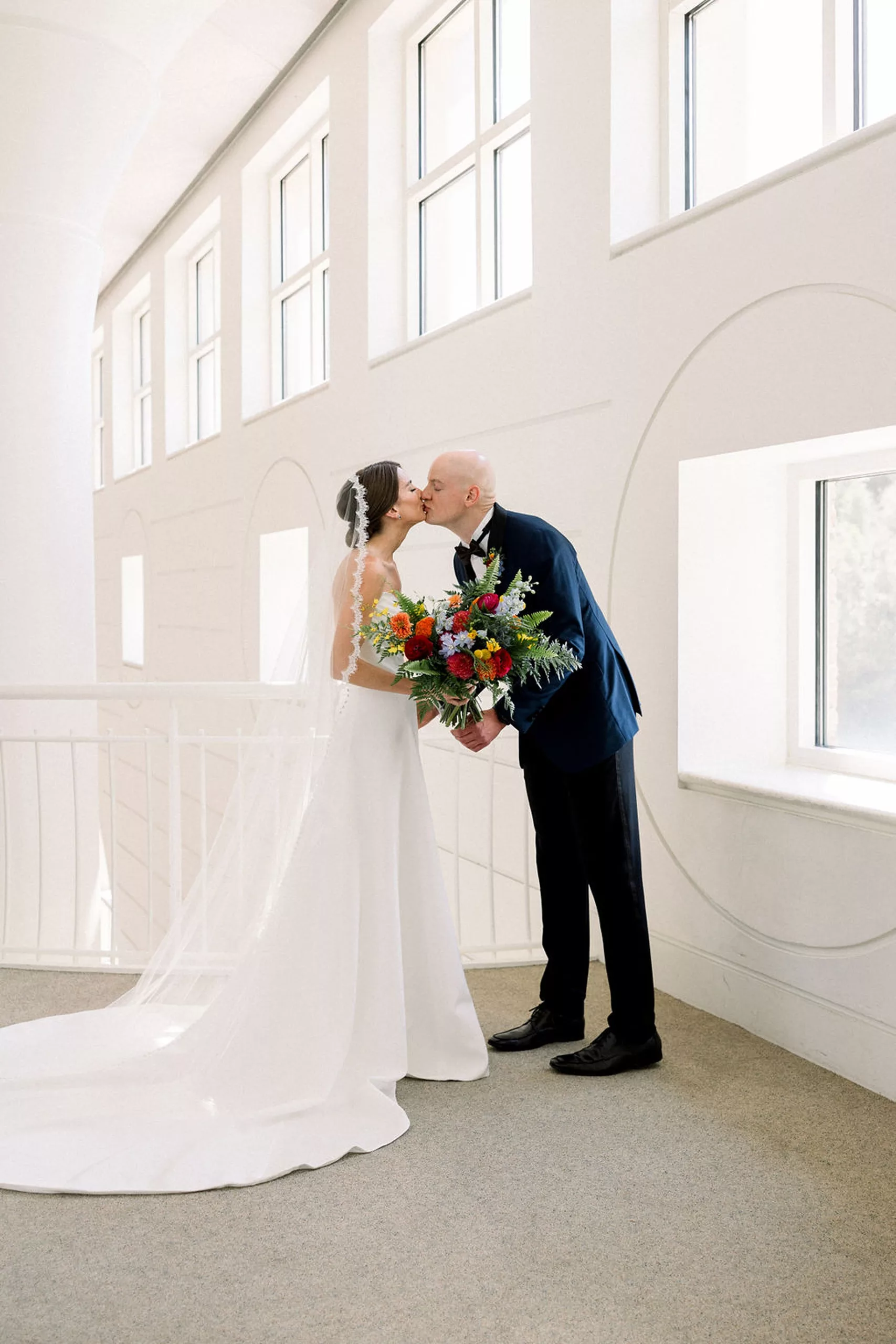 Newlyweds kiss while standing by large windows in a museum