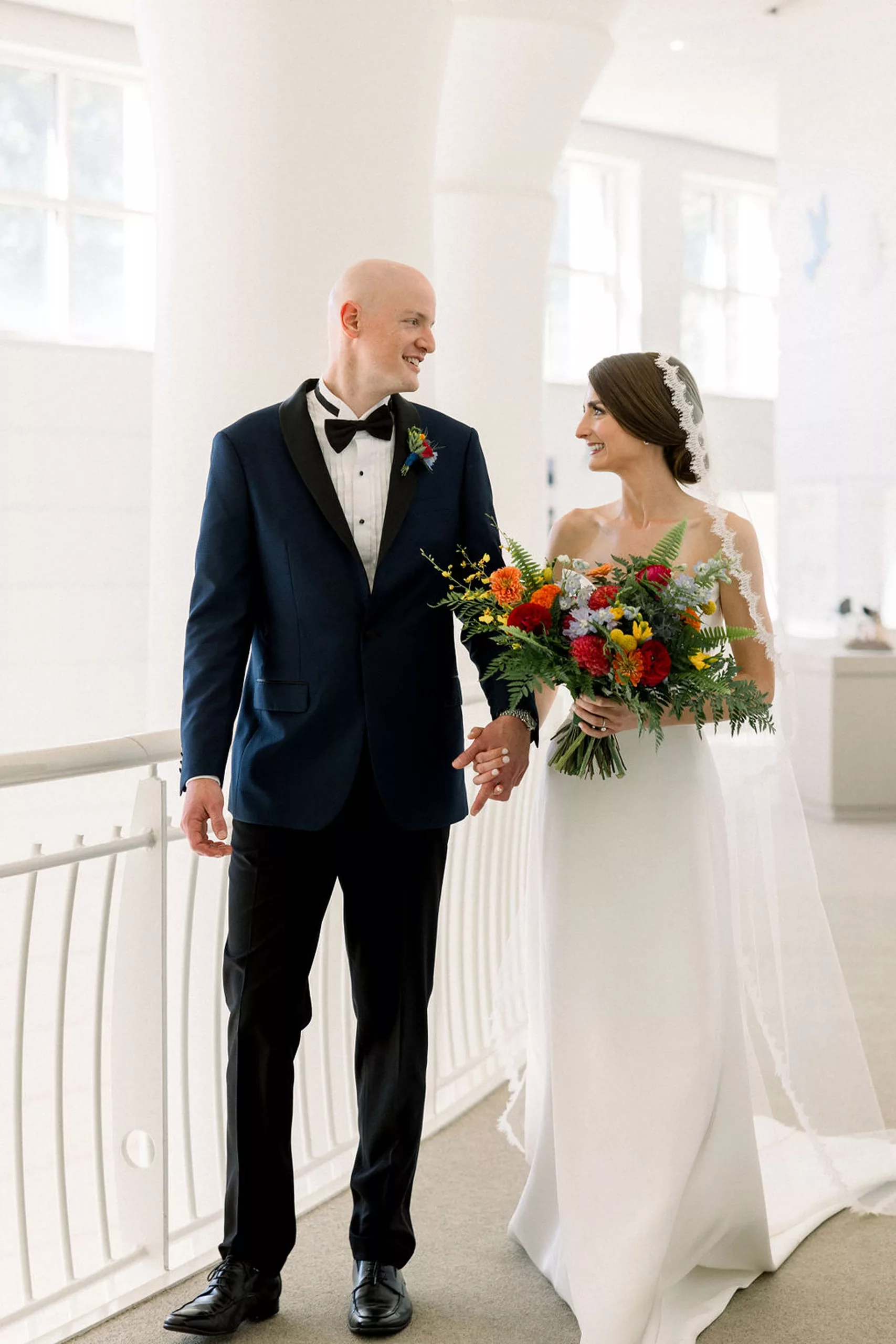 NEwlyweds walk hand in hand while looking at each other through a museum lobby by a railing
