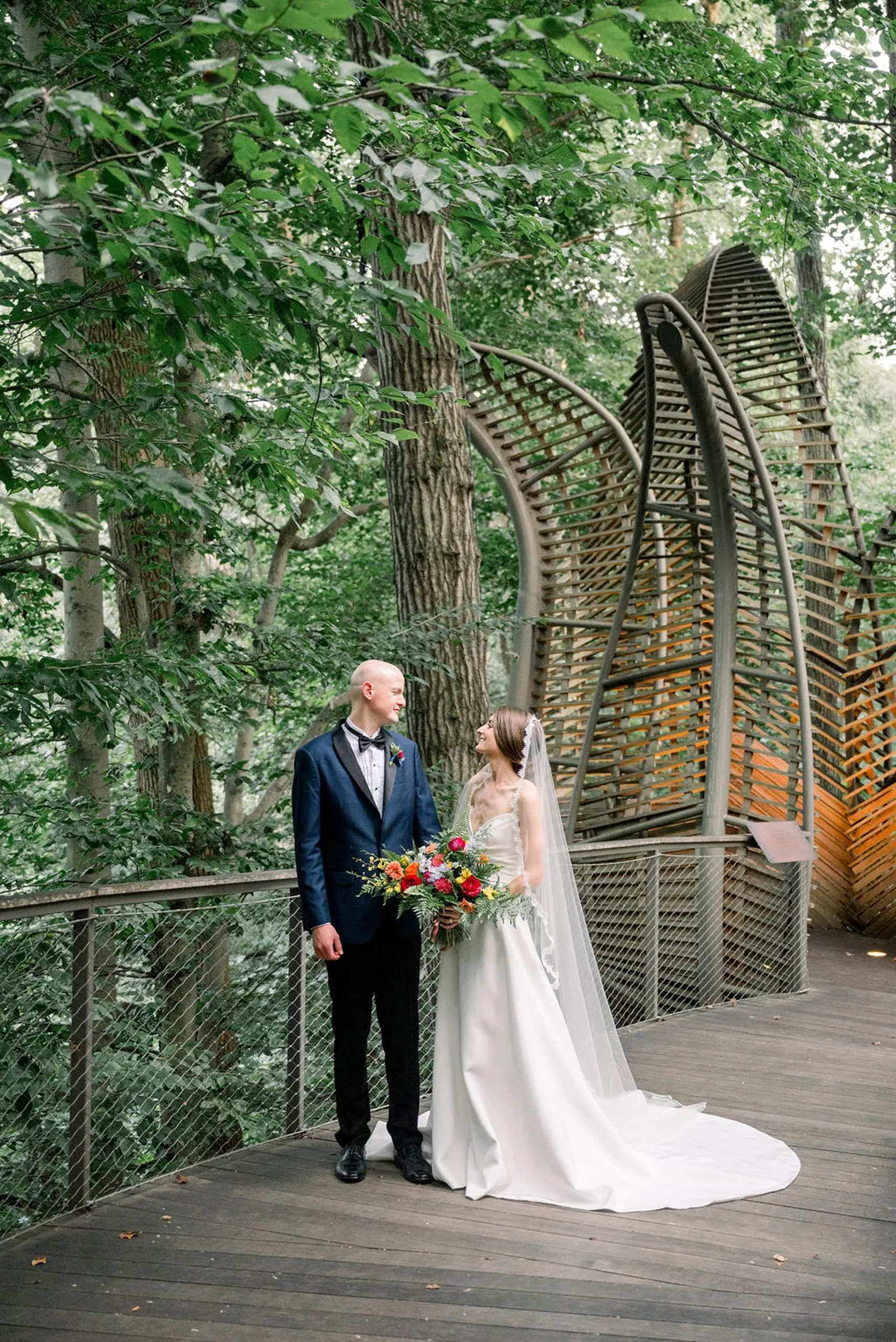 Newlyweds stand on a boardwalk in a forest looking at each other at their jurassic park wedding