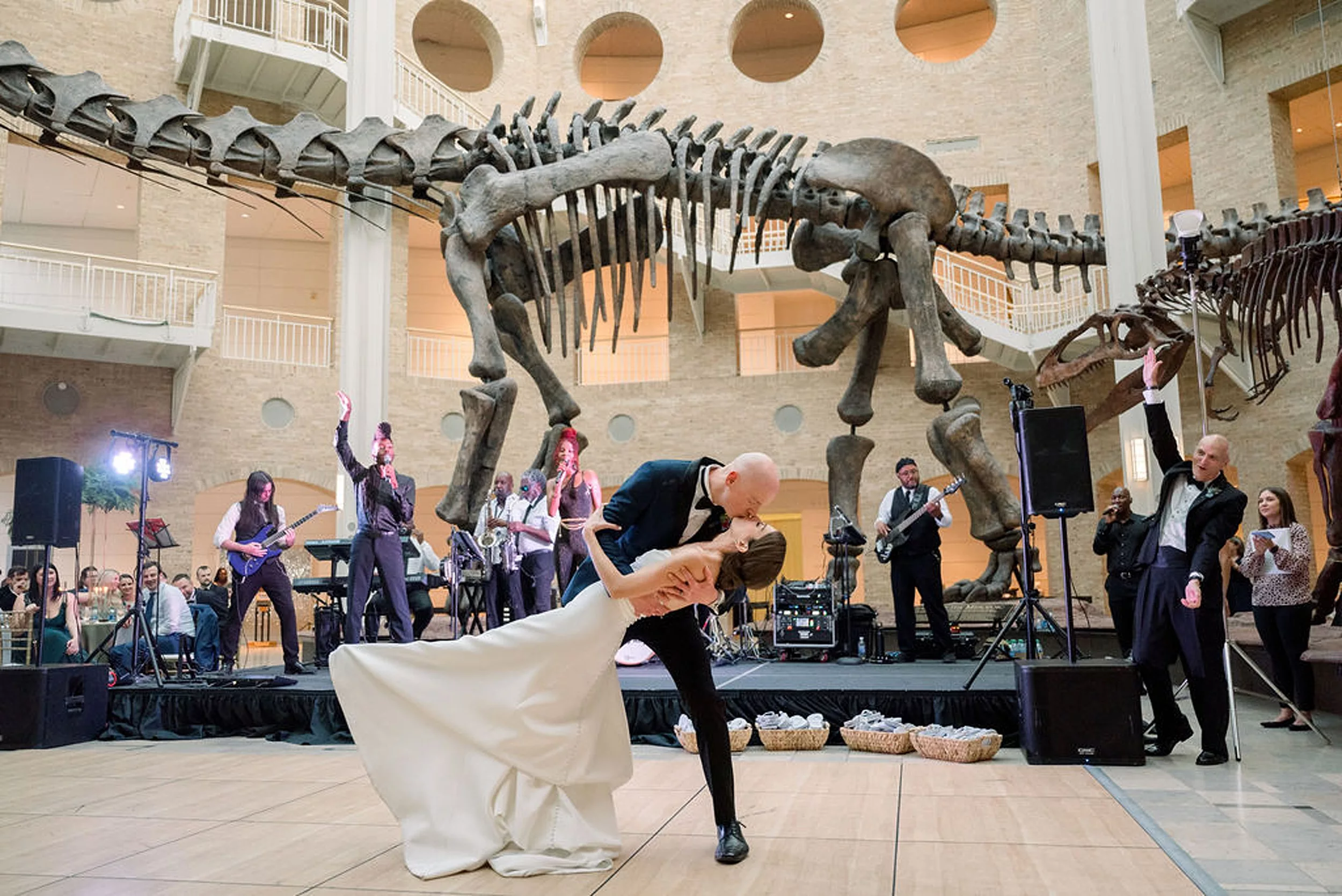 A groom dips his bride and kisses her as their band plays during their first dance in front of a dinosaur skeleton at their jurassic park wedding reception