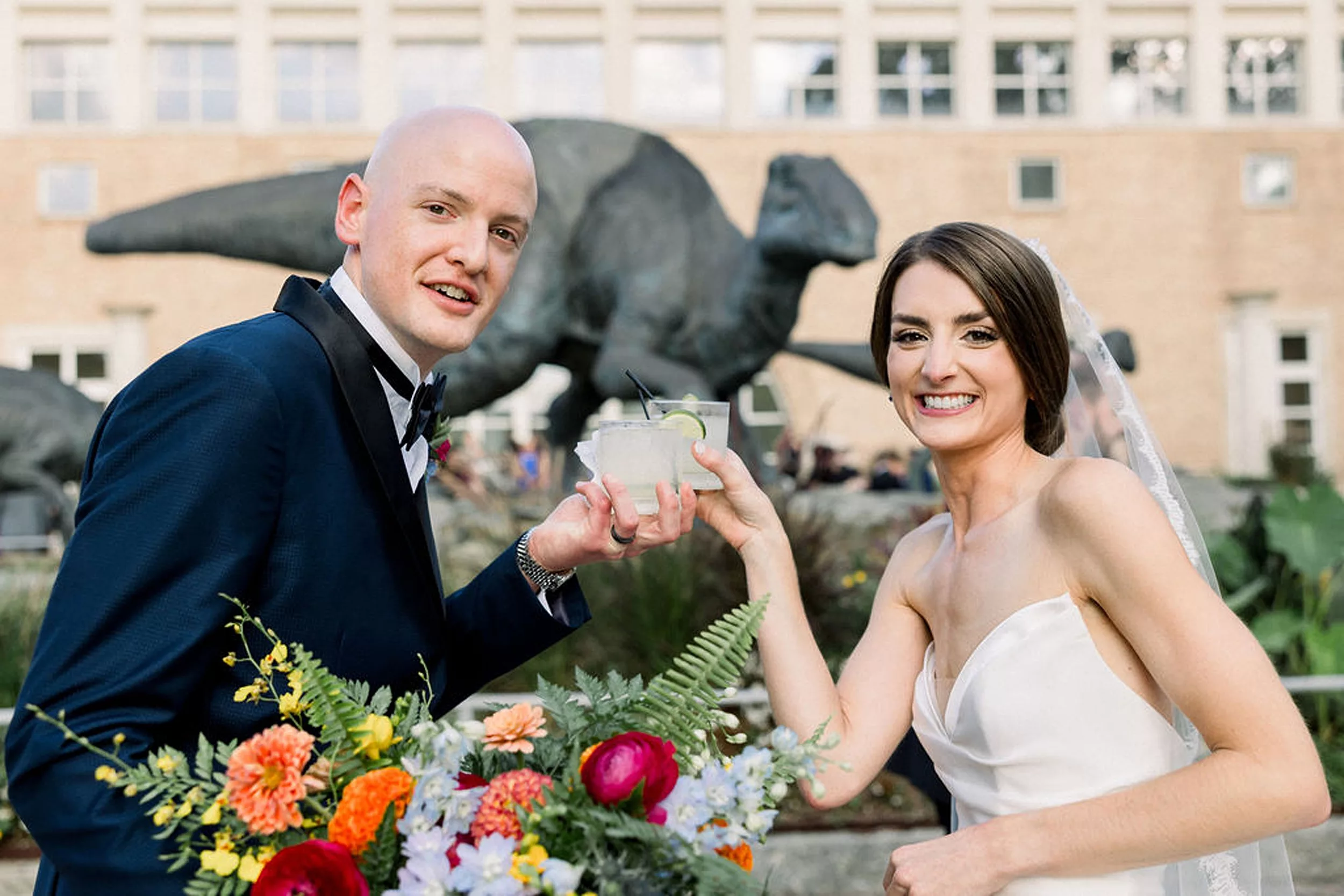 Newlyweds cheers their drinks while standing together in front of a dinosaur garden at their jurassic park wedding