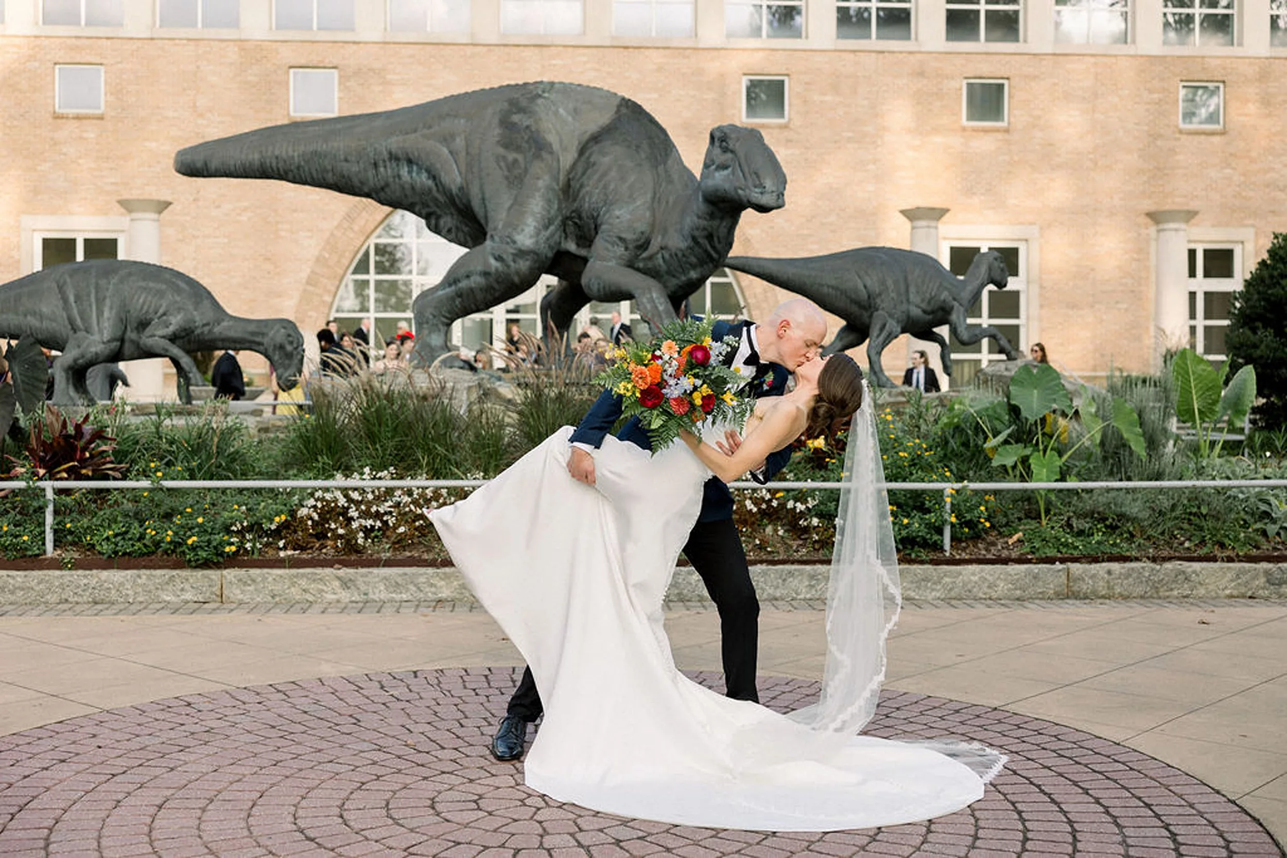 A groom dips and kisses his bride while standing in a dinosaur garden at their jurassic park wedding
