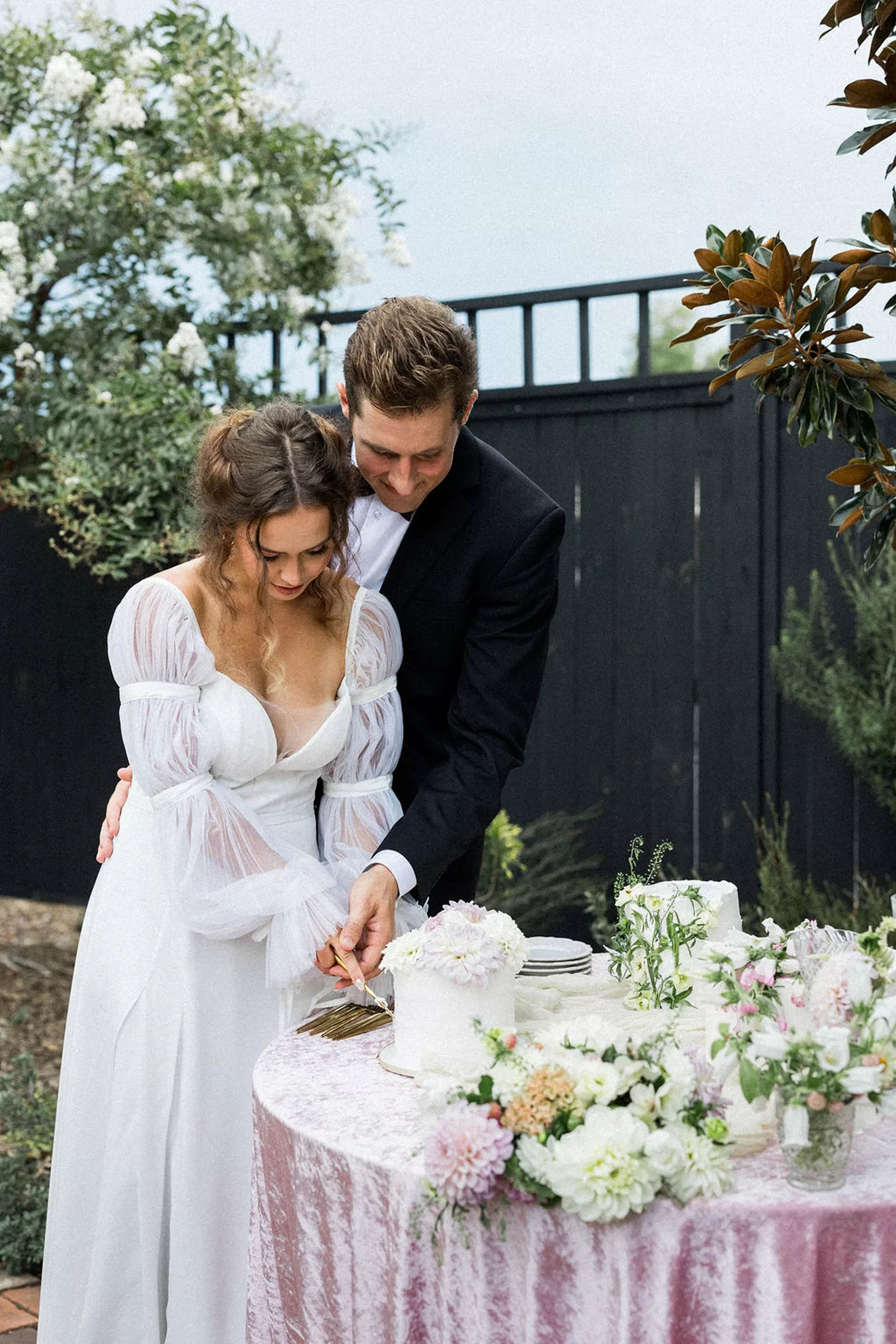 Newlyweds cut their cake together at a pink linen flower covered table in a garden at an lillian gardens wedding