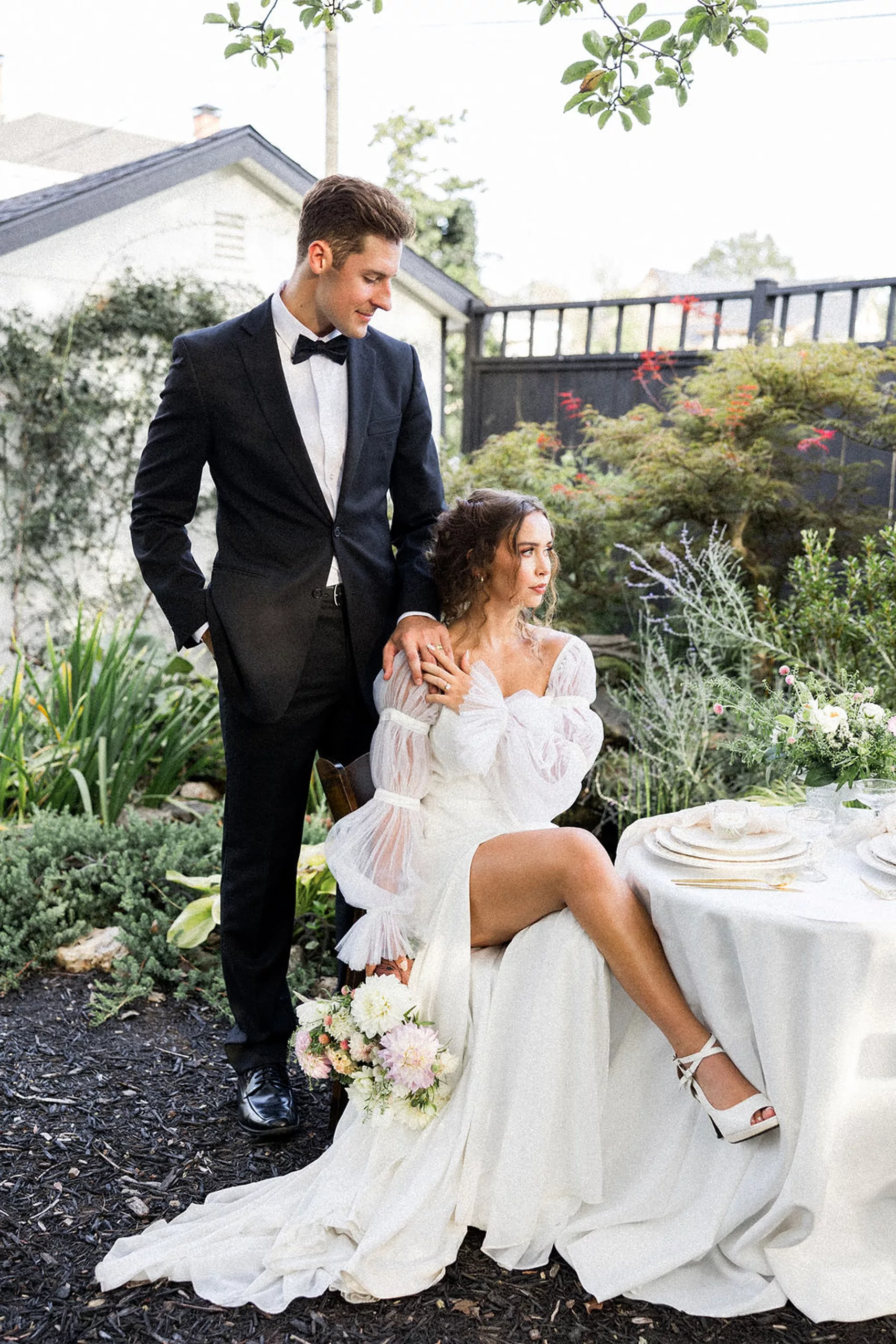 A bride sits in a chair with legs crossed as her husband stands behind her with a hand on her shoulder in a garden