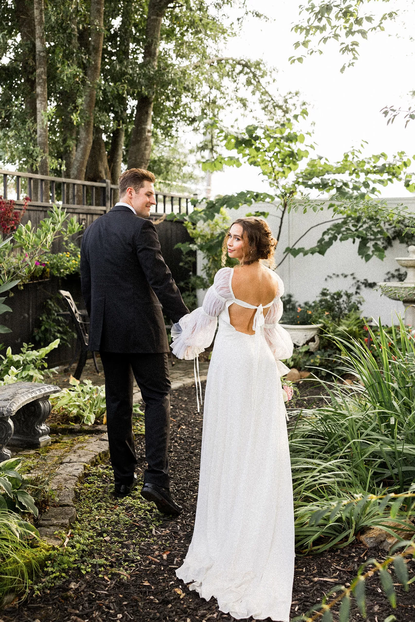 A groom leads his bride by the hand as they walk through a garden by a stone bench and bird bath
