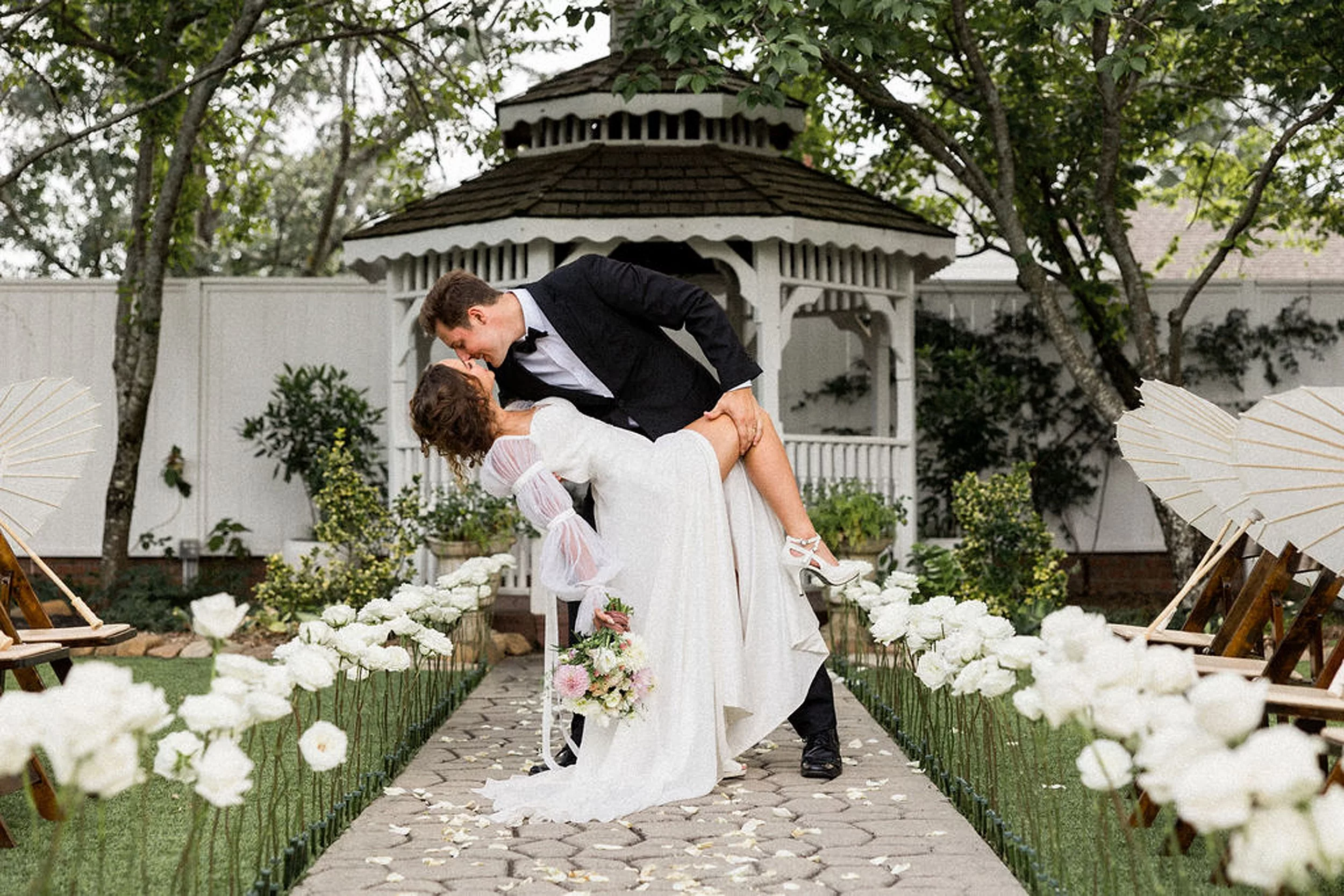 A groom kisses and dips his bride in the stone aisle leading to a gazebo lined with white roses