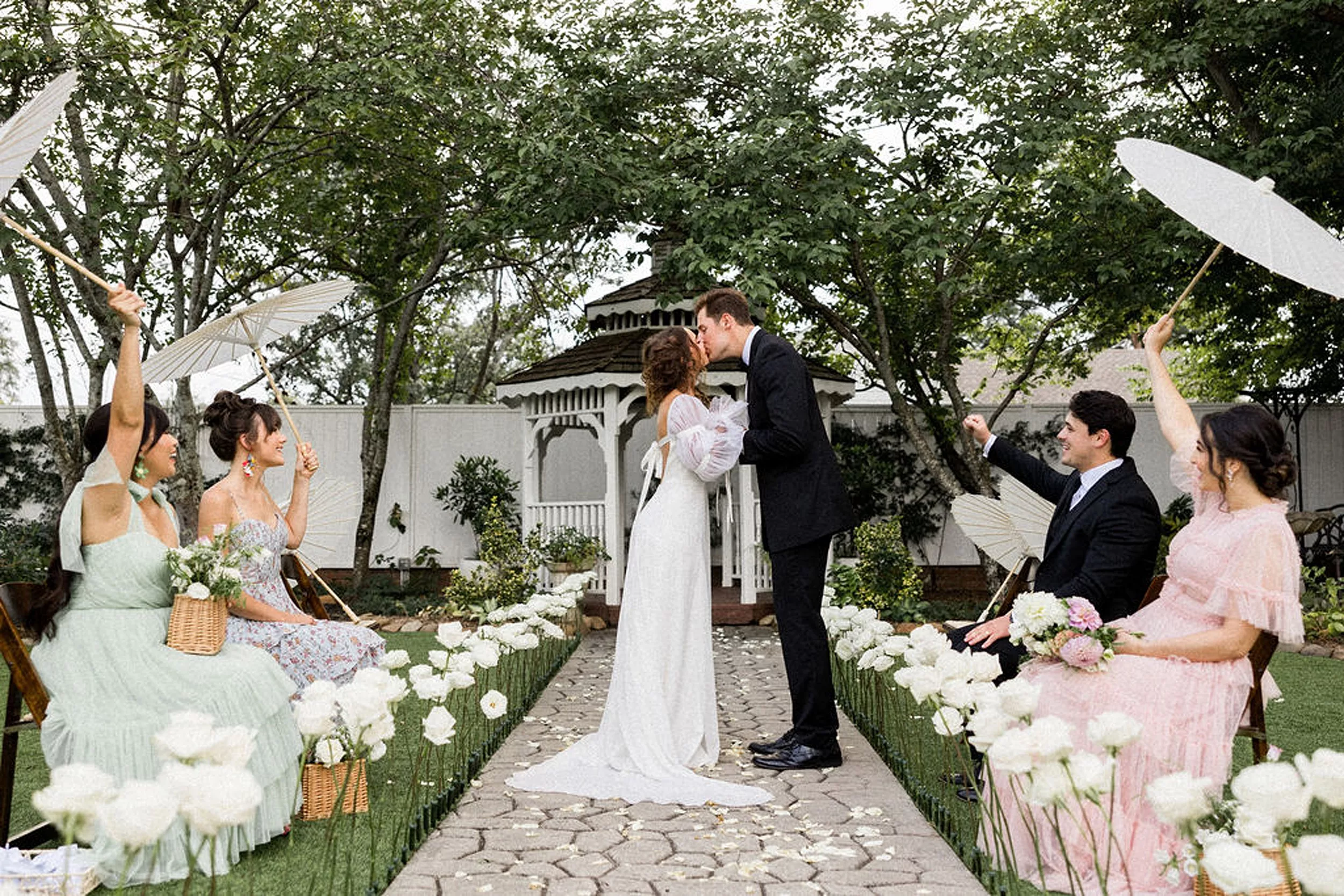 Newlyweds kiss while standing in the stone aisle of a garden wedding ceremony as their guests cheer while holding umbrellas