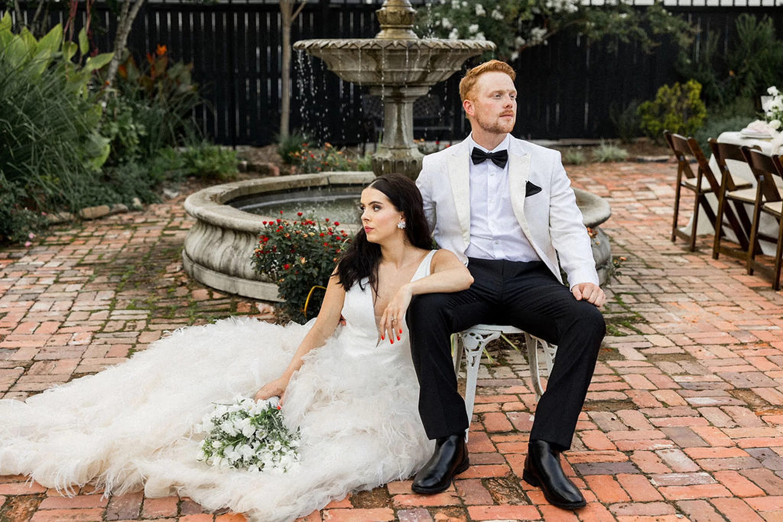 Newlyweds sit in a chair and on the brick pavers by the fountain in a garden