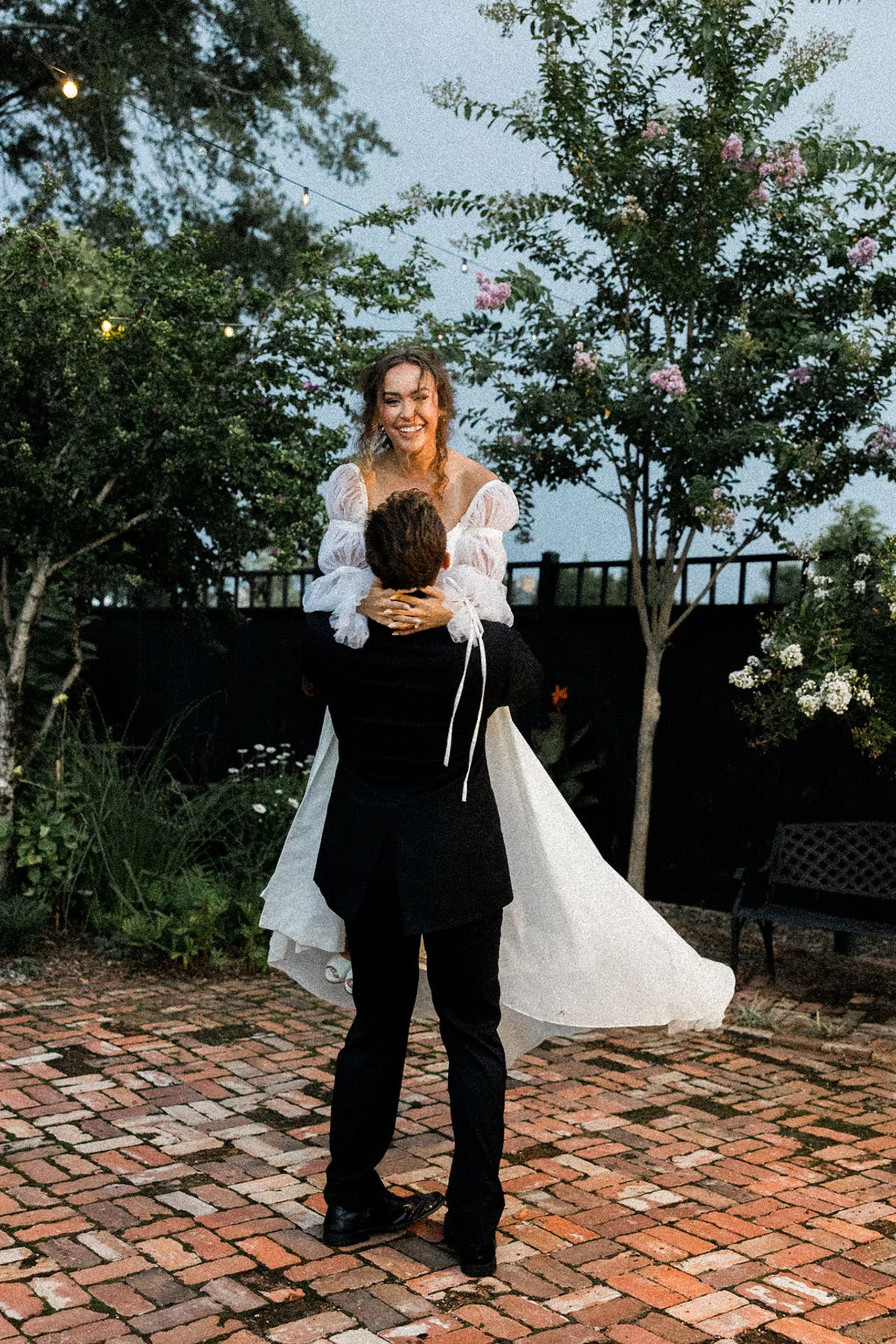 A groom in a black suit dances and lifts his bride while they dance on the brick patio outside the lillian gardens wedding venue
