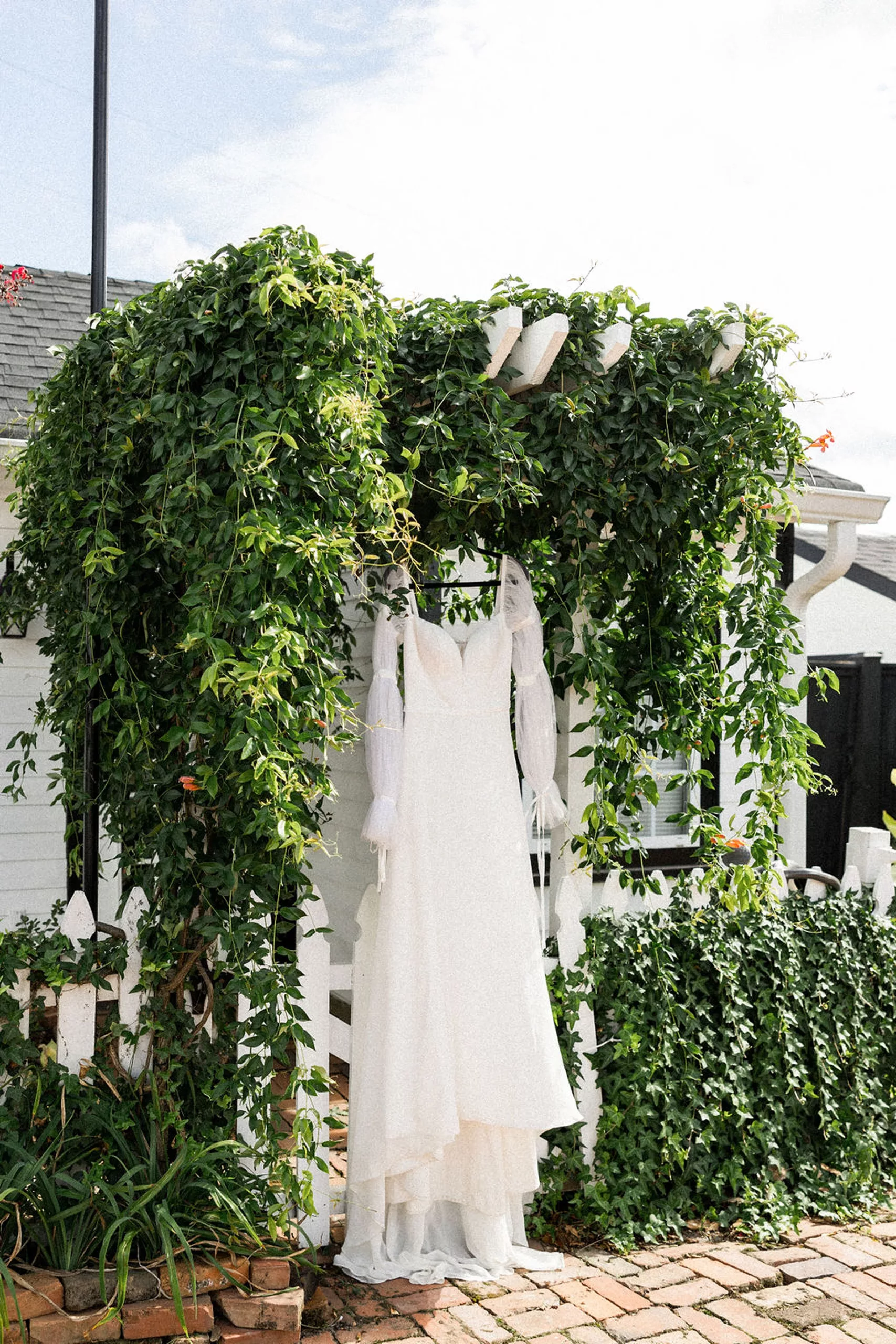 A bride's dress hangs from a vine covered pergola on a brick path in the lillian gardens wedding venue garden