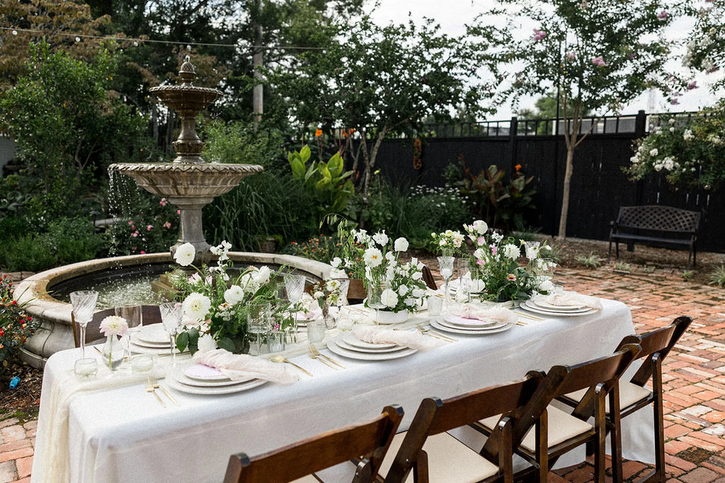 Details of an outdoor wedding reception table setup with wooden chairs and white florals by the fountain on a brick path through the lillian gardens wedding venue