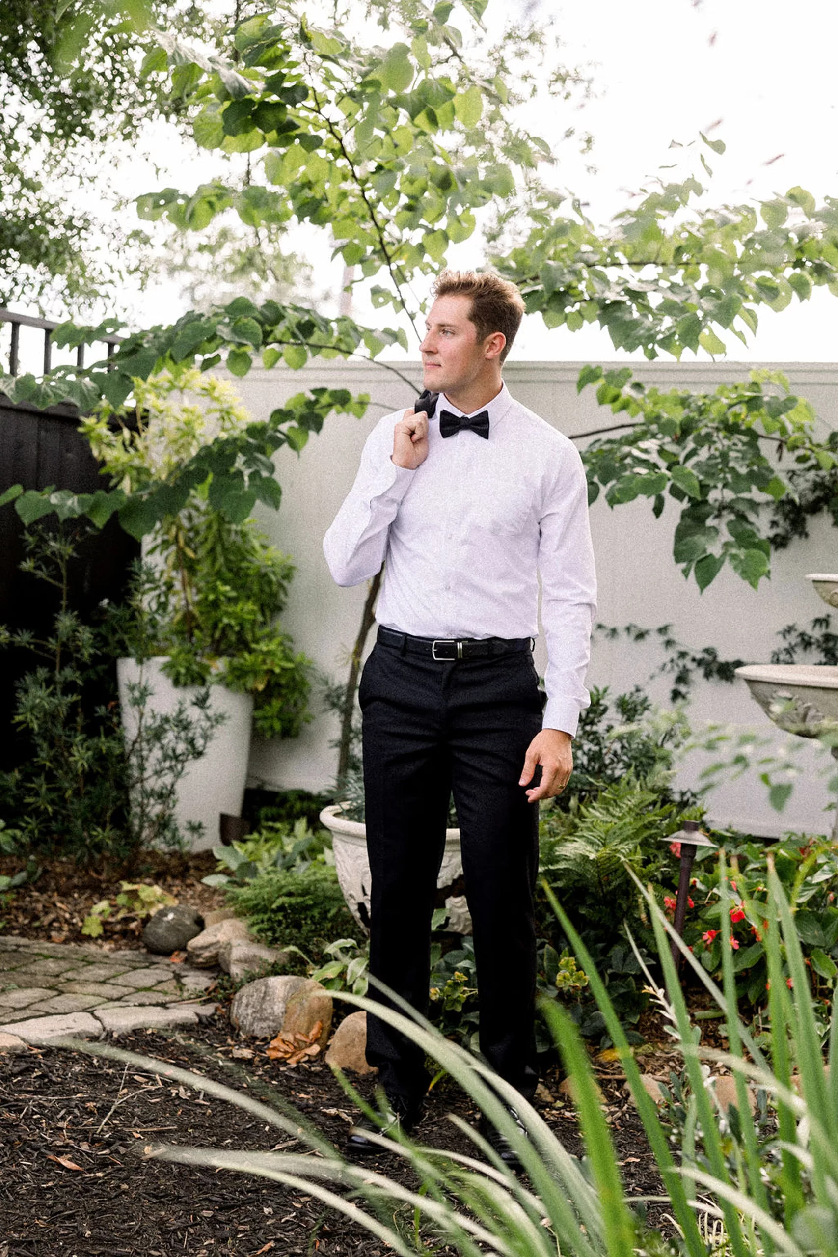 A groom holds his suit jacket over his shoulder while standing in the garden by a bird bath in the lillian gardens wedding venue