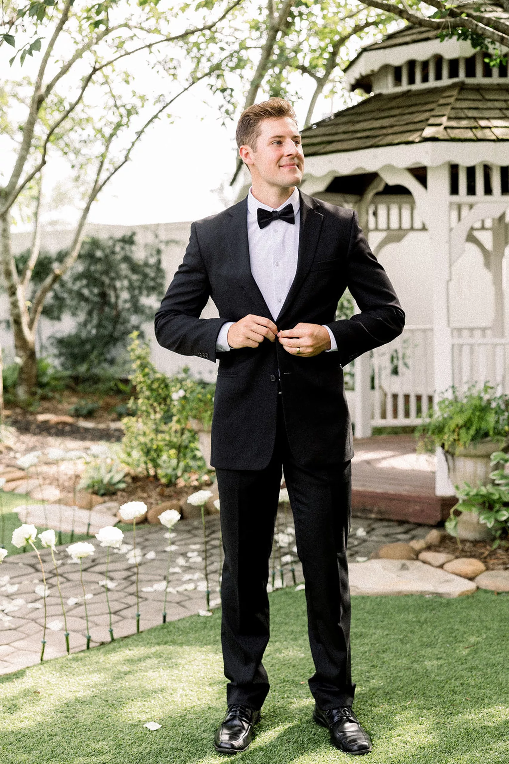 A groom in a black suit adjusts his buttons while looking off into the lillian gardens wedding ceremony location