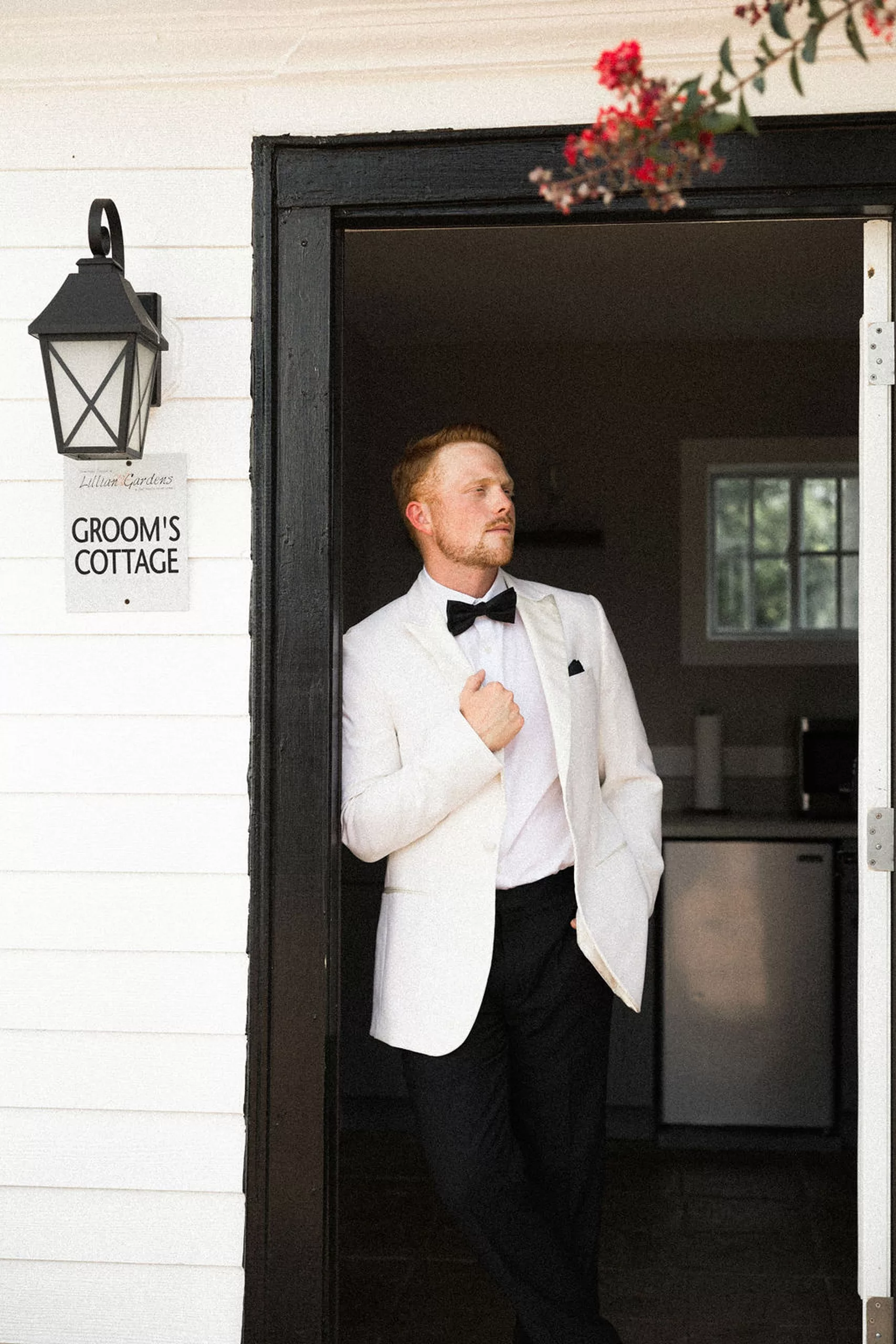 A groom holds his lapel of his white suit jacket at the lillian gardens wedding venue groom's cottage door