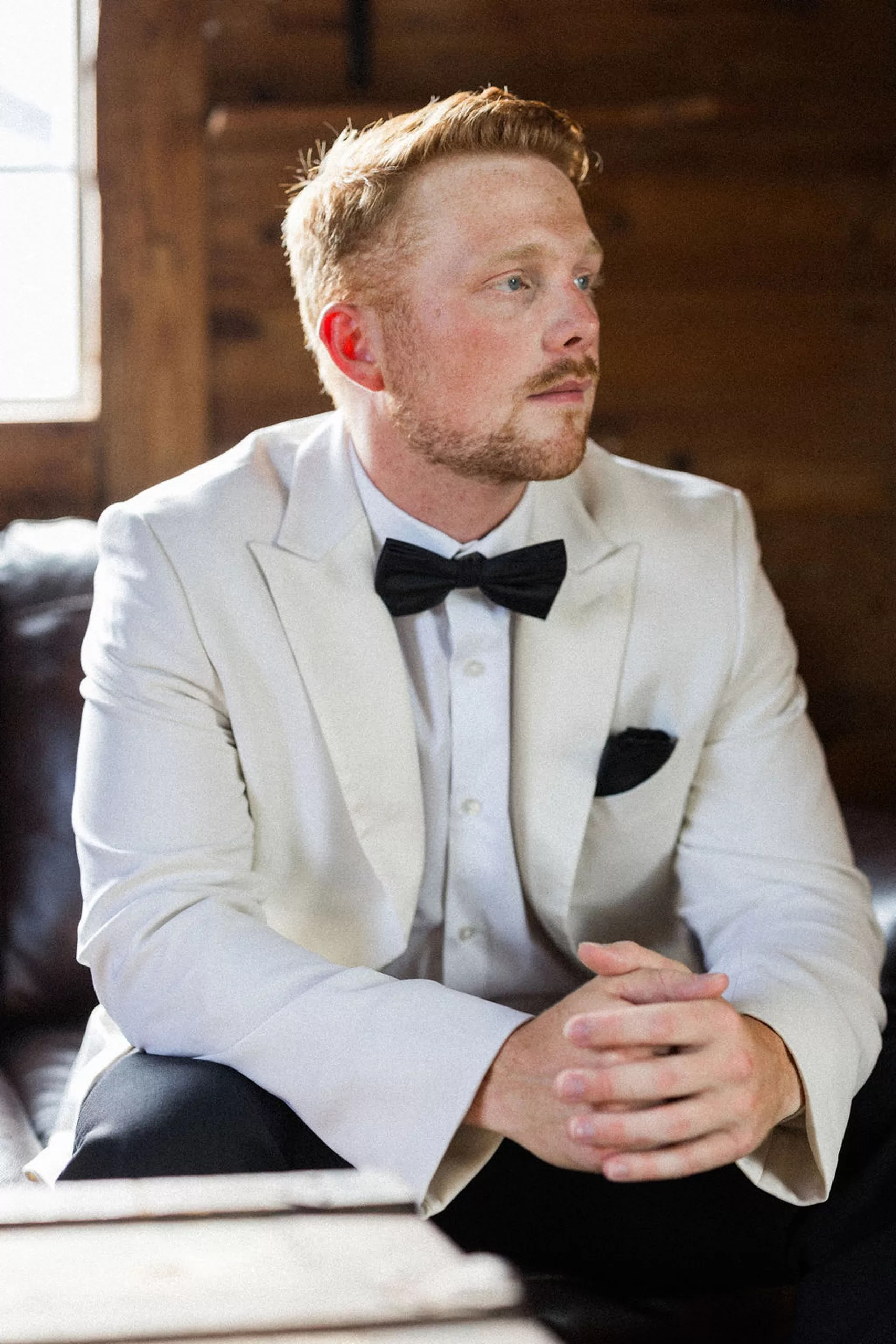 A groom in a white suit jacket and black pants sits on a leather chair with hands together looking over his shoulder at an lillian gardens wedding