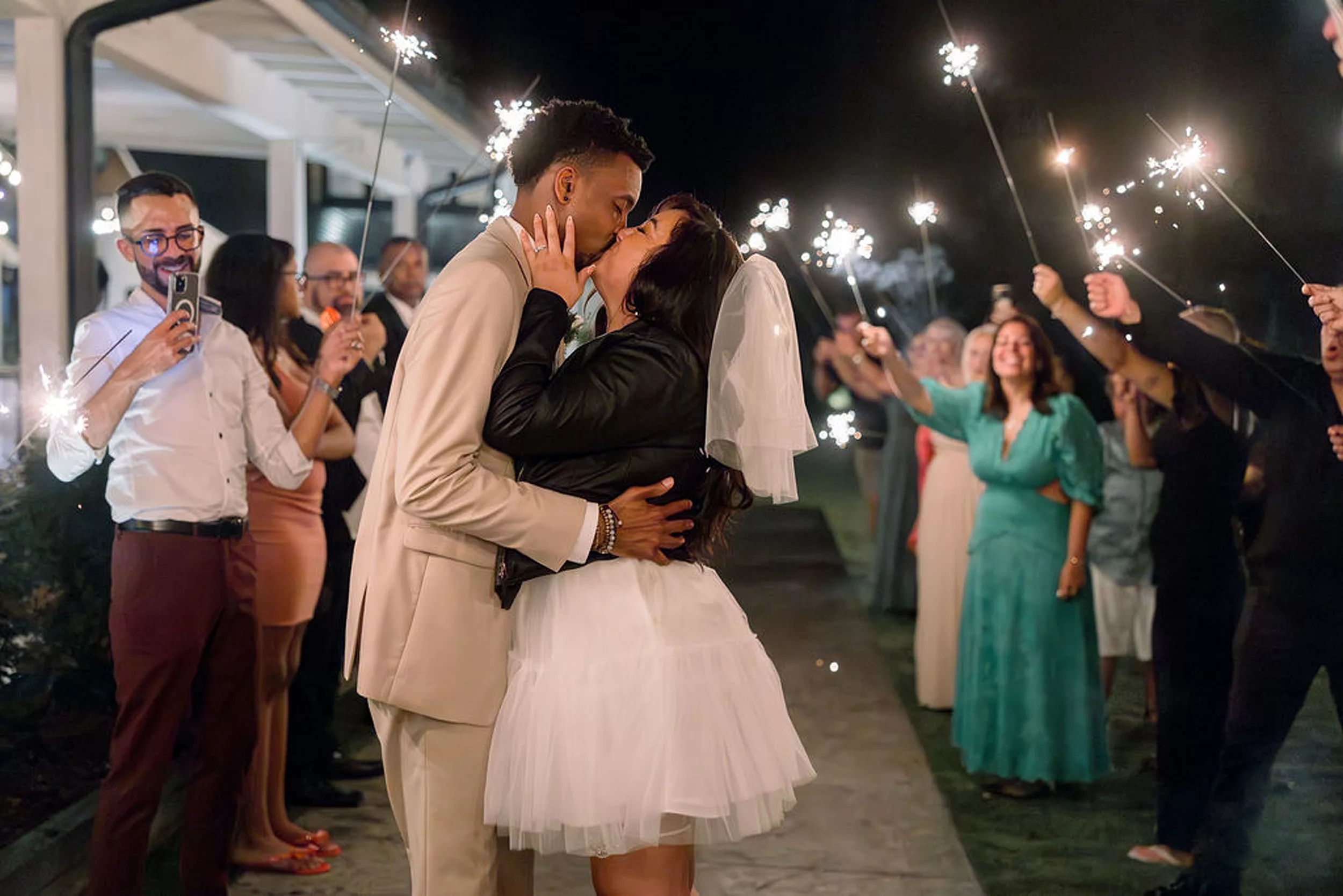 Newlyweds kiss under a shower of sparklers held up by their wedding guests on a sidewalk