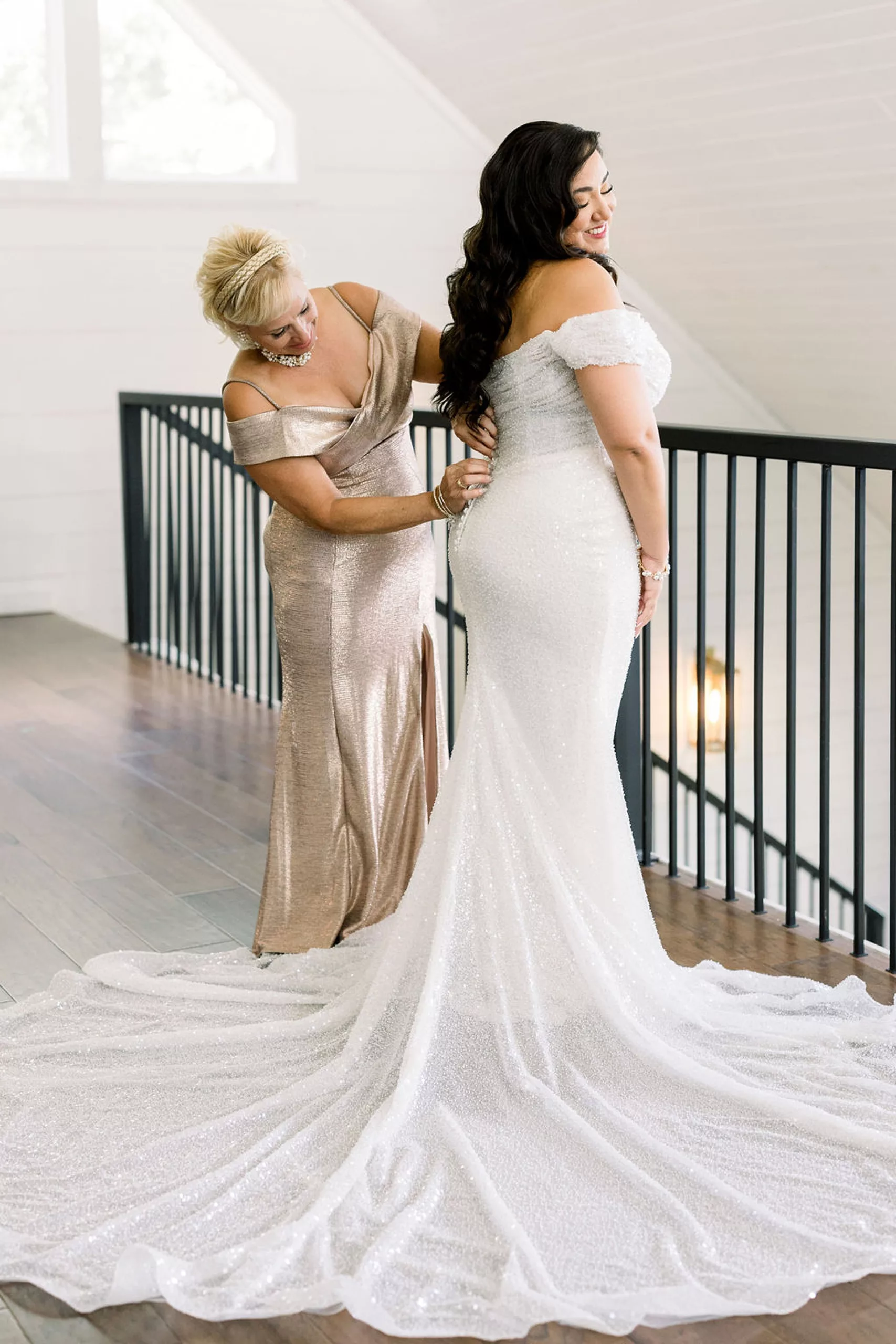 A bride stands on a balcony with her dress spread as her mom helps button the back