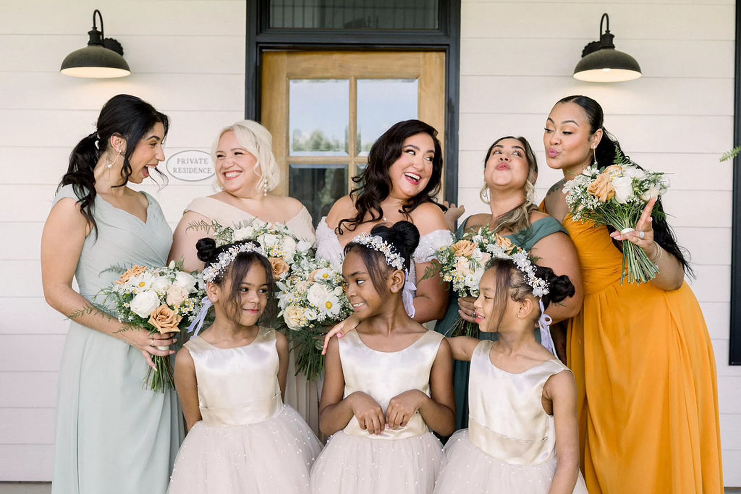 A bride laughs and plays with her bridesmaids and four flower girls while standing on a porch