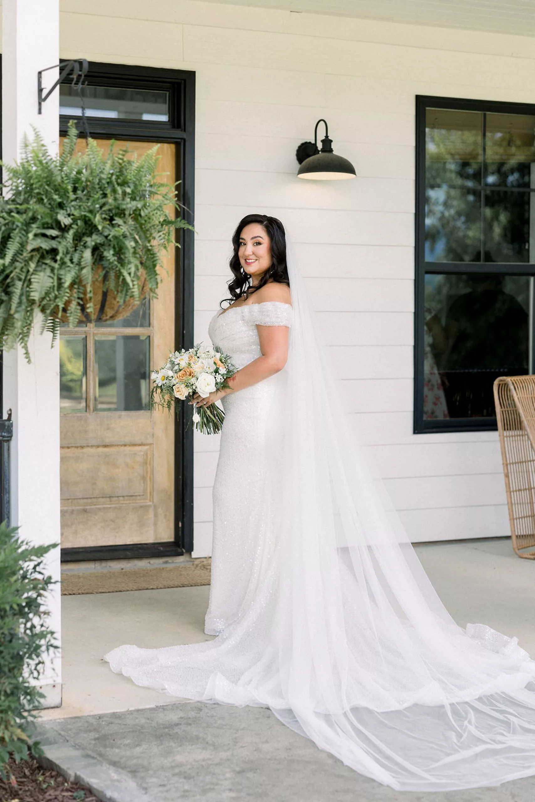 A bride stands on a porch with her train spread holding her white bouquet