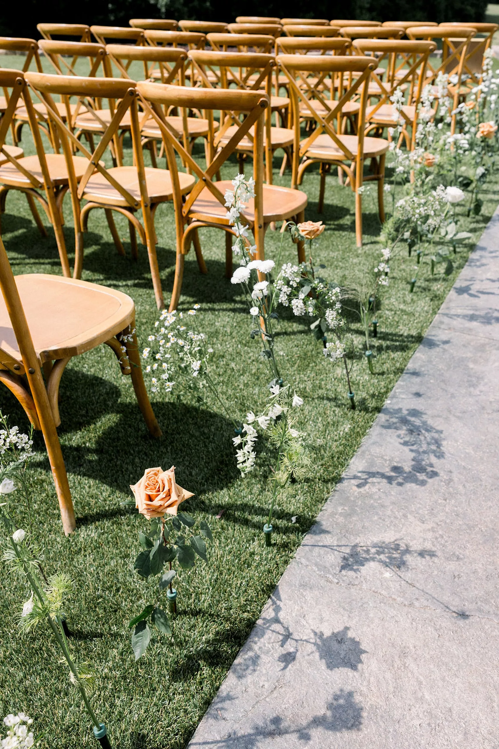 Details of wooden chairs set up for a wedding ceremony along a cement path through grass lined with white and pink flowers at meadows at mossy creek