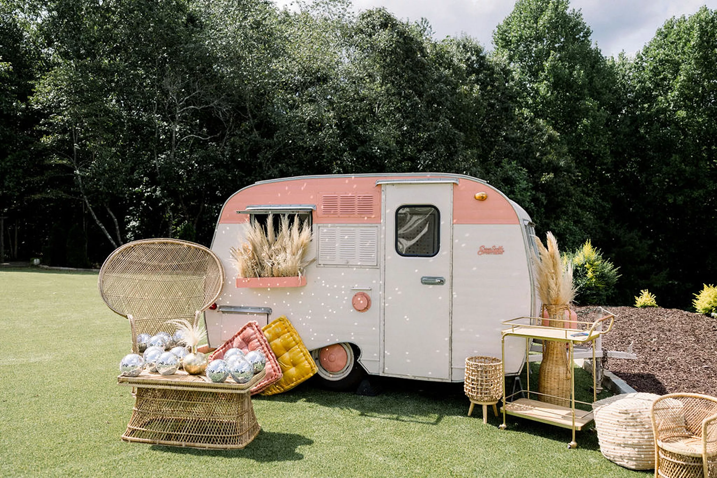 A small pink trailer sits with boho and wicker themed decorations in a field at a meadows at mossy creek wedding