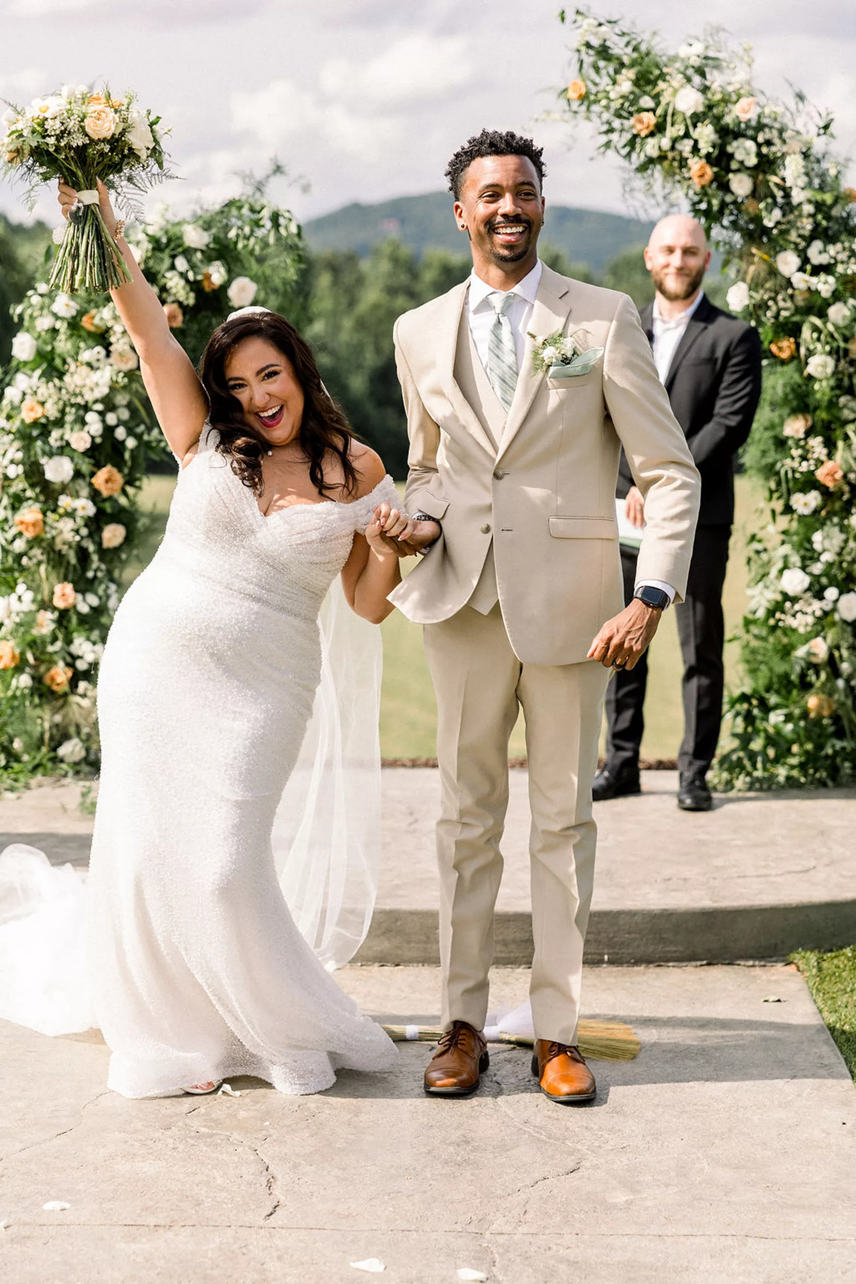 A bride raises her bouquet over her head in celebration while holding hands with her groom in a tan suit after jumping over the broom for their meadows at mossy creek wedding ceremony