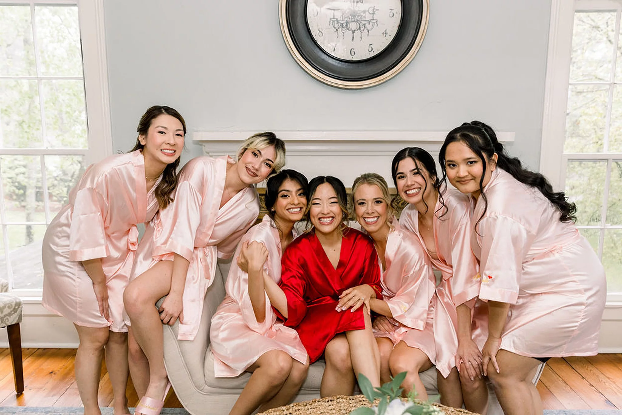 A bride laughs and sits on a couch with her six bridesmaids in pink robes while she wears a red robe