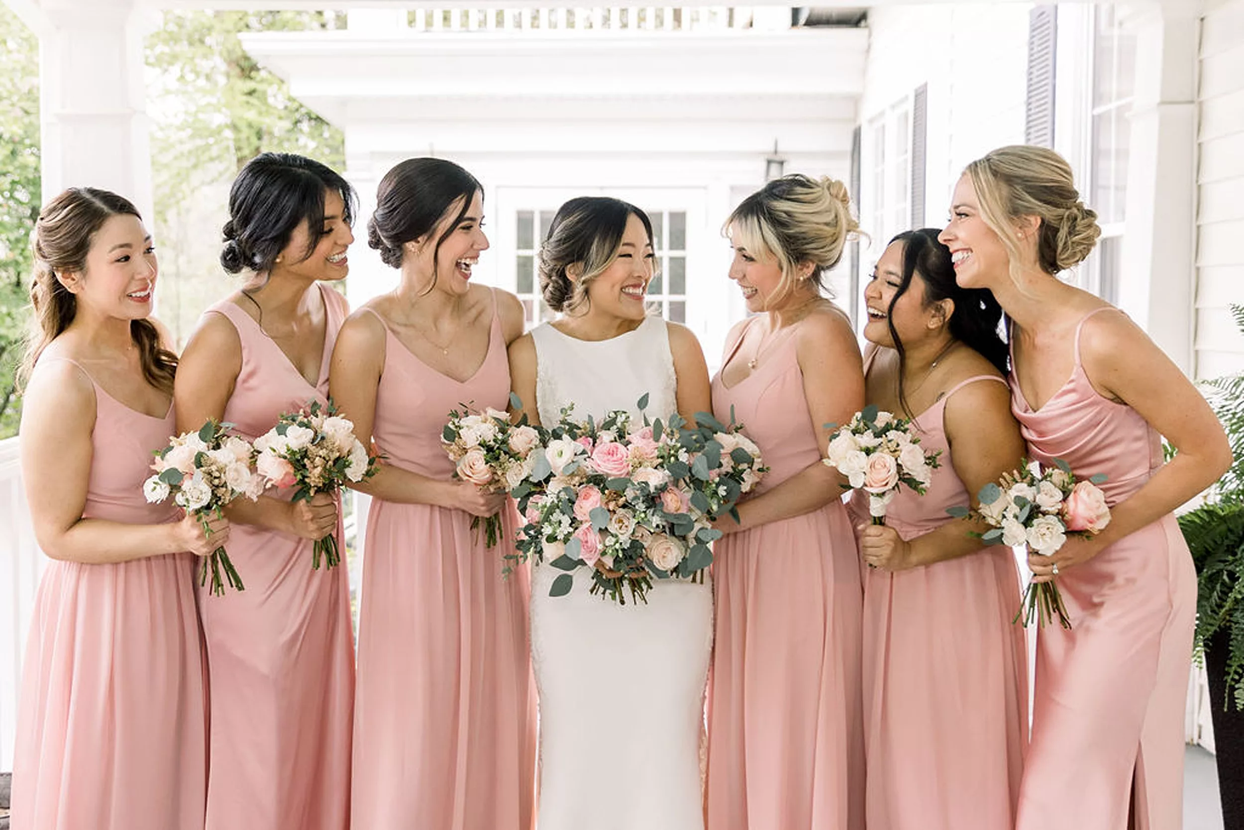 A bride smiles and laughs with her bridesmaids in pink dresses on a porch