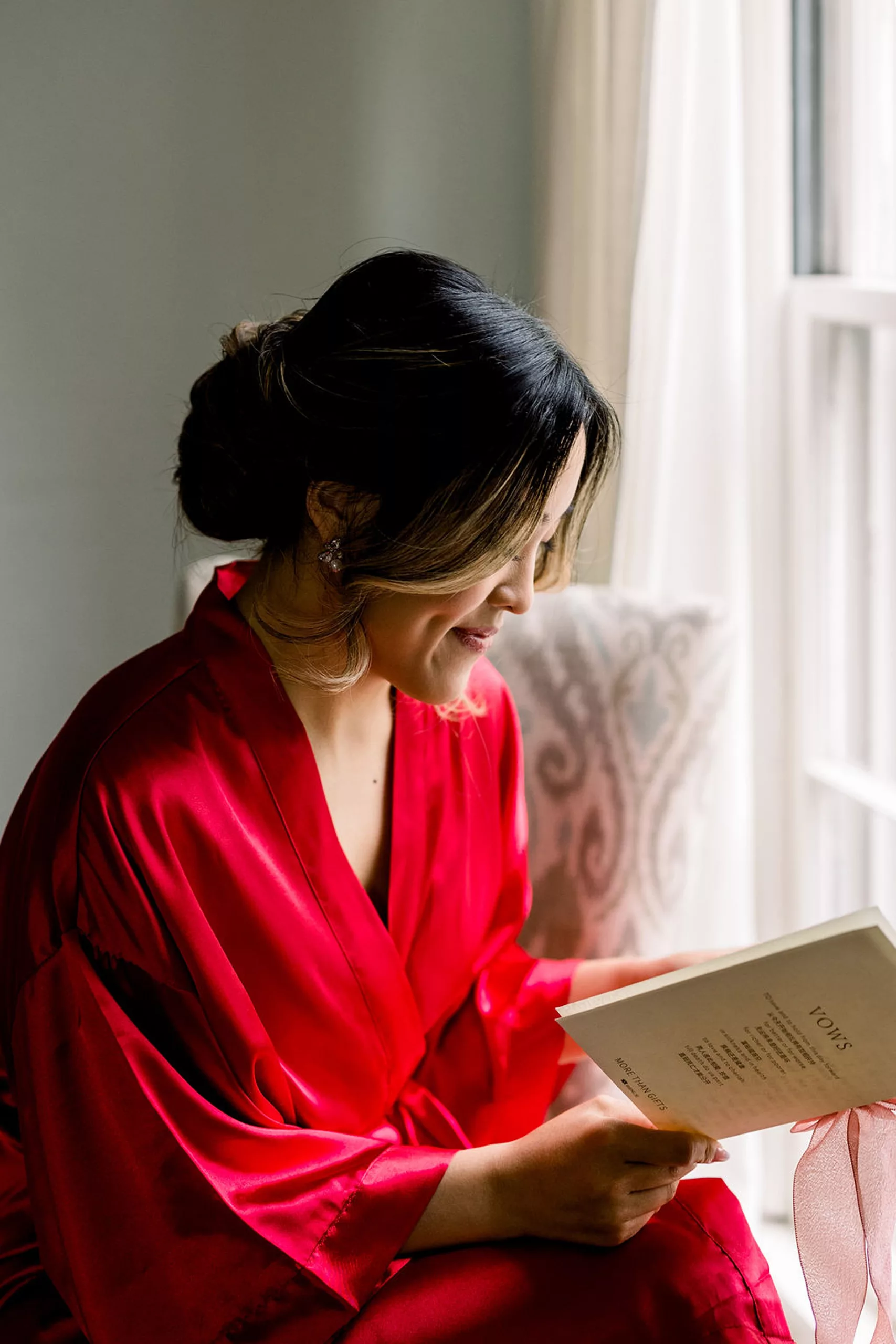 A bride in a red robe sits in a window reading her vows before getting ready