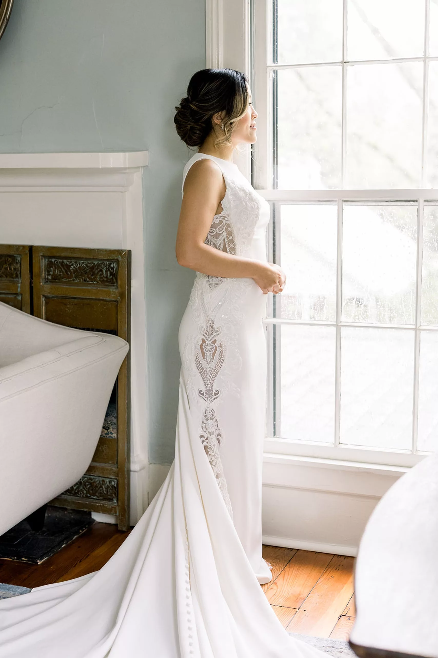 A bride stands in a window in her lace embroidered dress