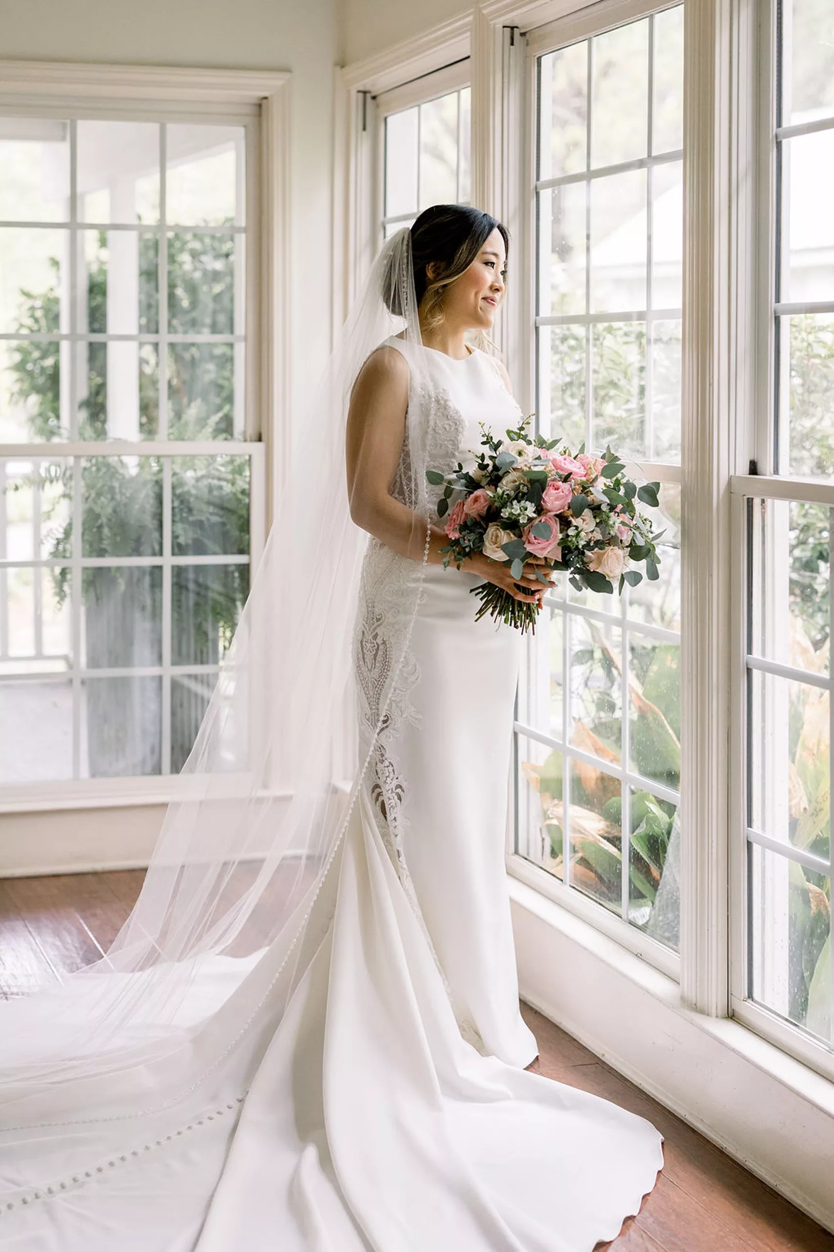 A bride stands in a sun room smiling out the window while holding her pink and white bouquet