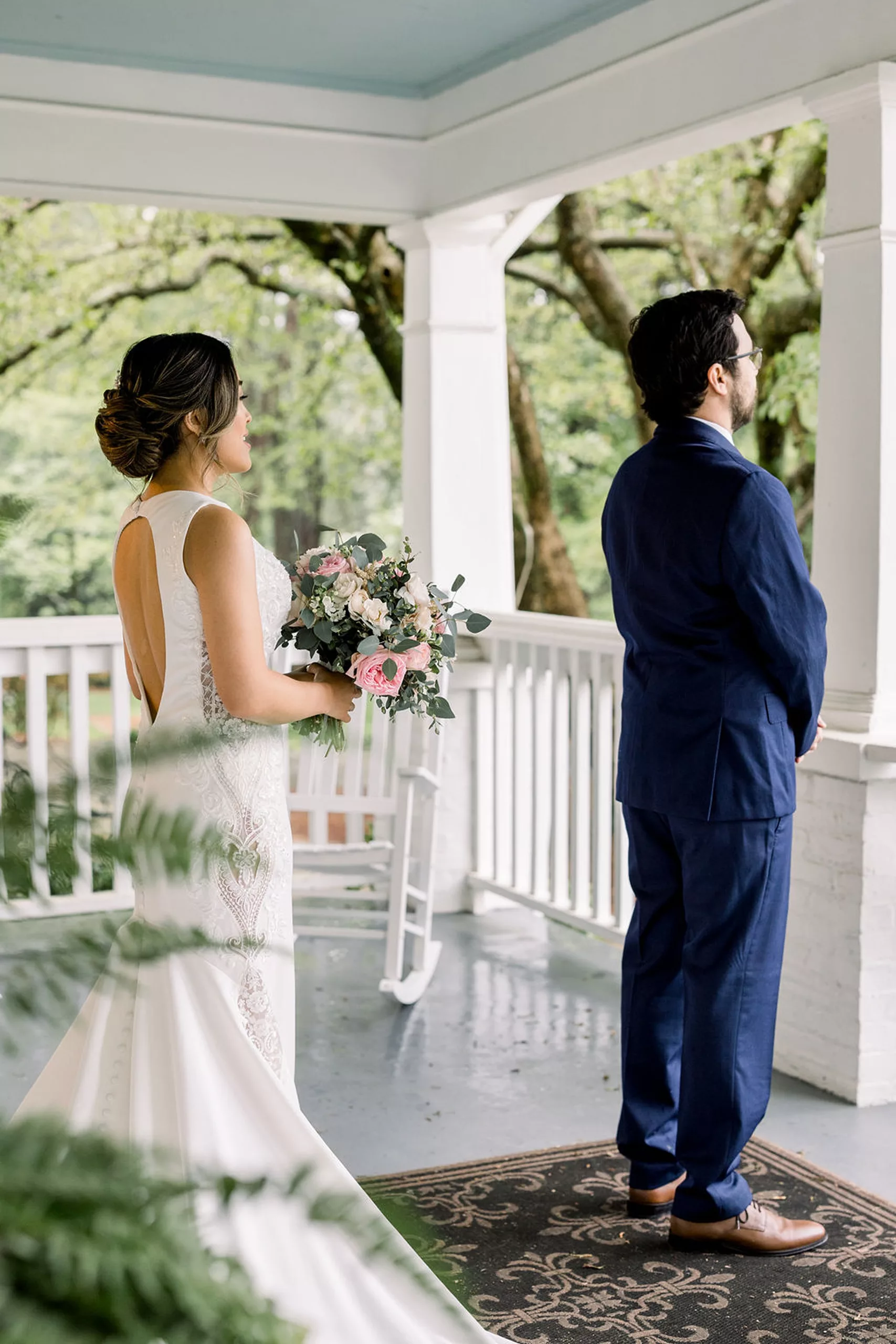 A groom stands facing away from his bride as she approaches for a first look on a porch