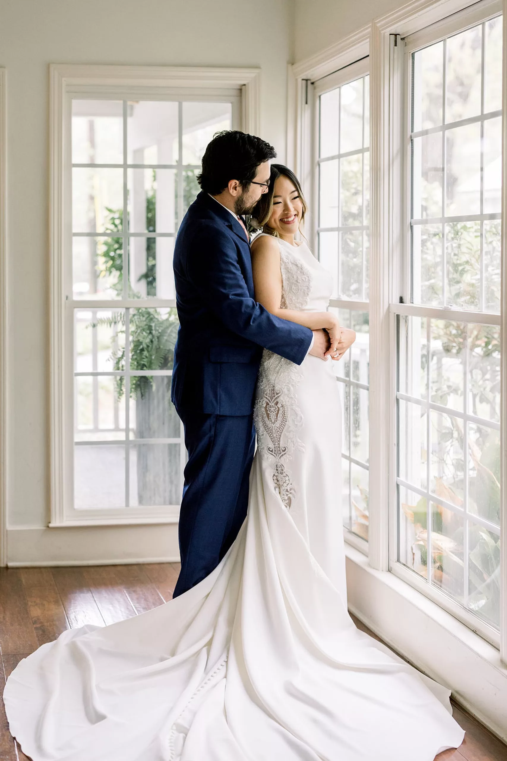 A bride leans into her husband in a blue suit as they snuggle in a large window with the train spread out