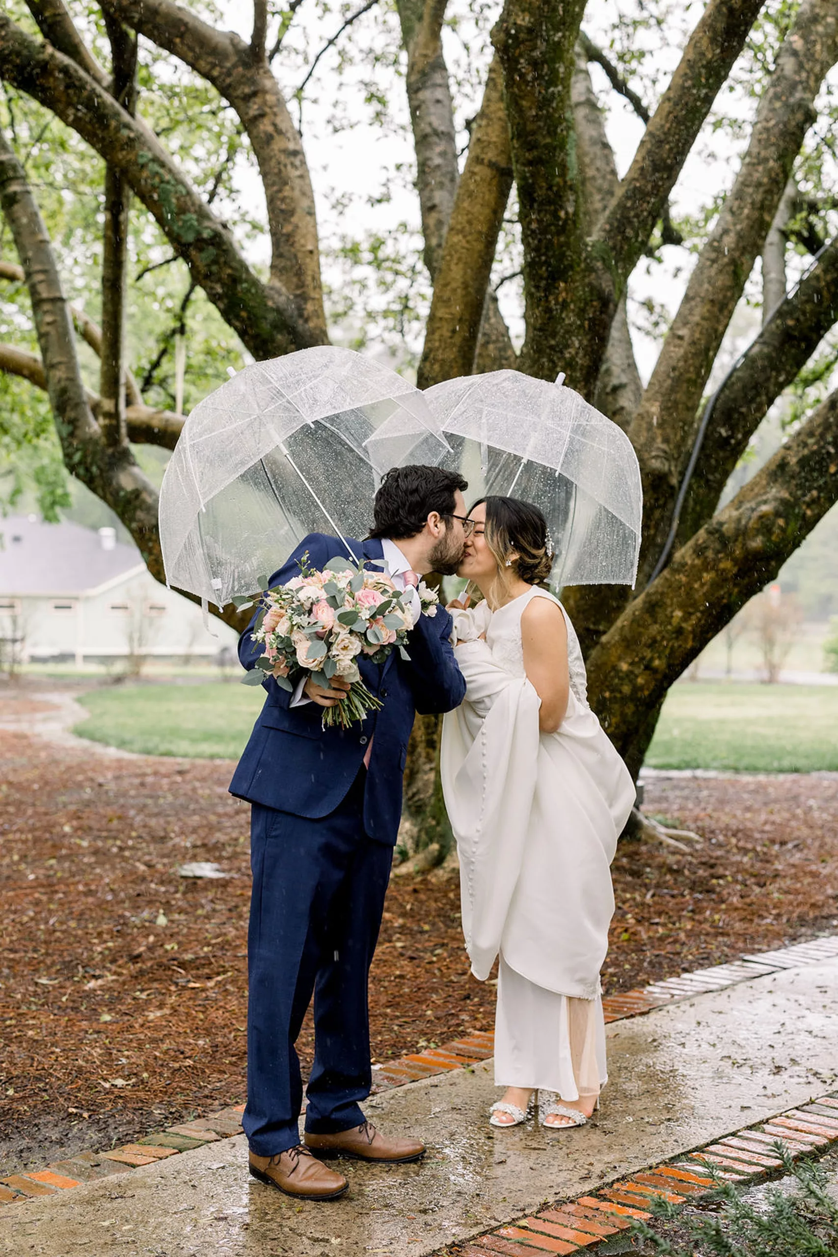 Newlyweds kiss under clear umbrellas on a raining day on a garden sidewalk lined with bricks