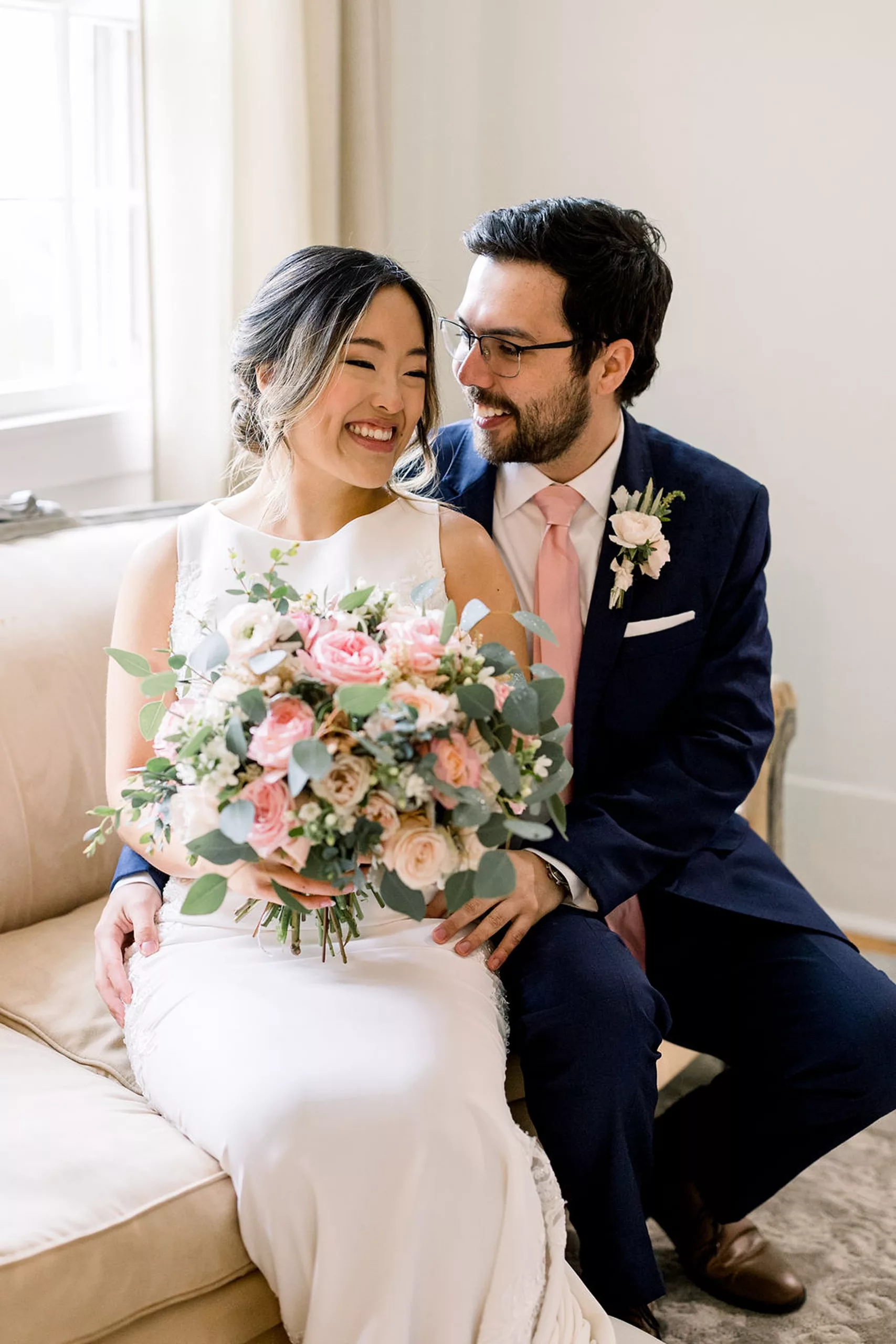 A groom in a blue suit and pink tie sits and hugs his bride on a tan couch under a window