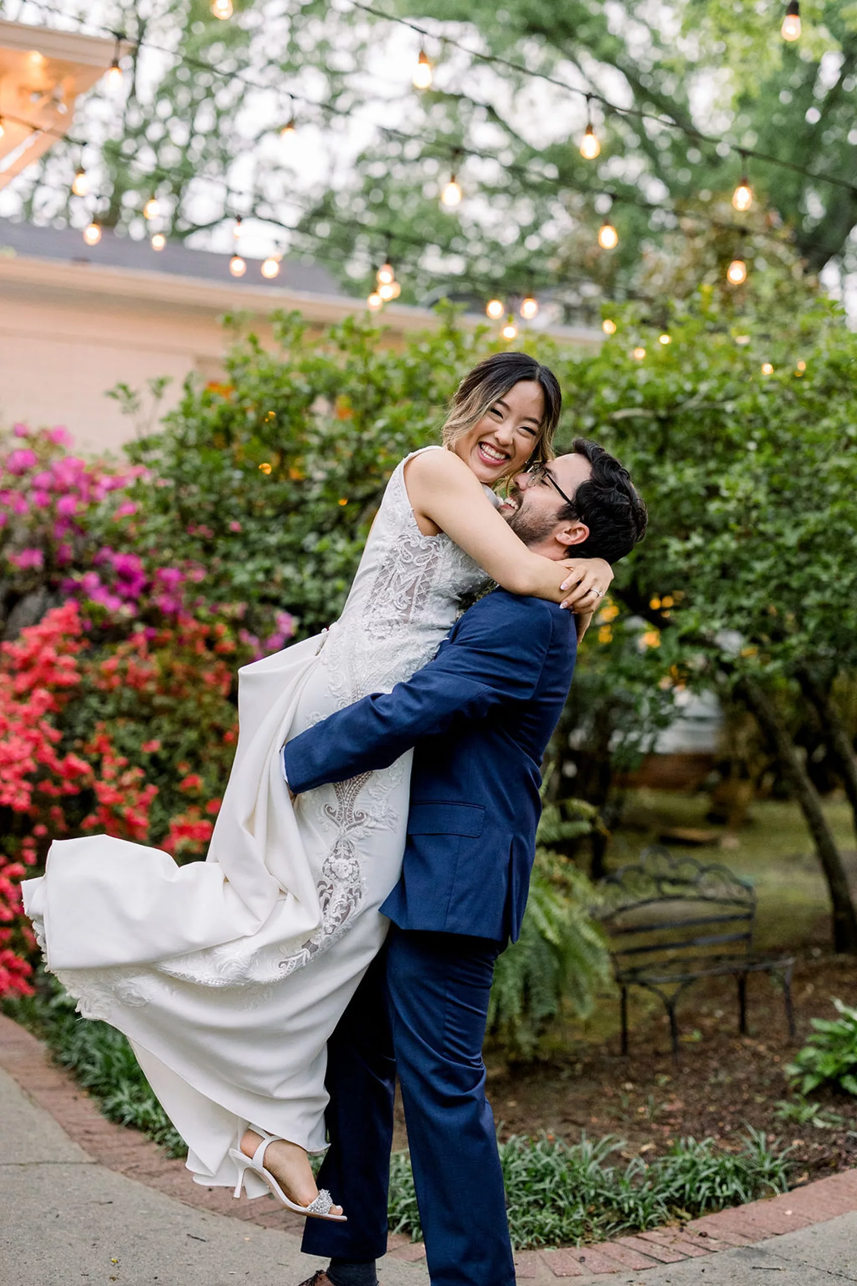 A groom in a blue suit lifts and laughs with his bride in a flower garden at a payne-corley house wedding