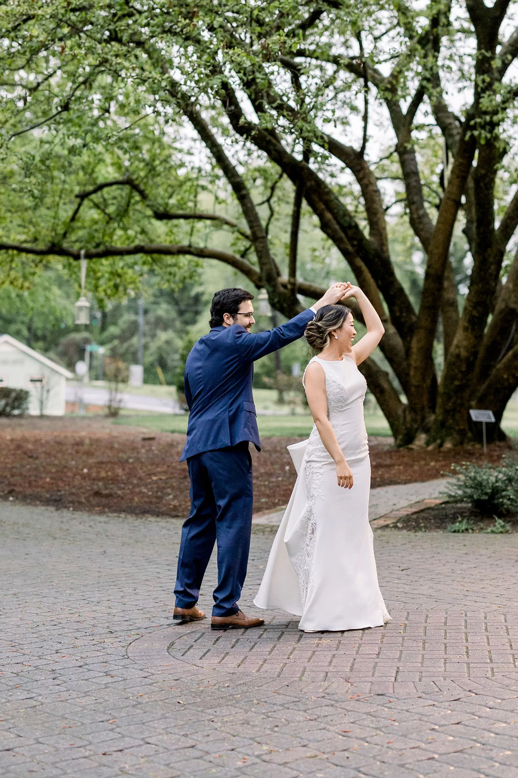 A groom in a blue suit twirls his bride on a stone patio under a large tree at the payne-corley house wedding venue