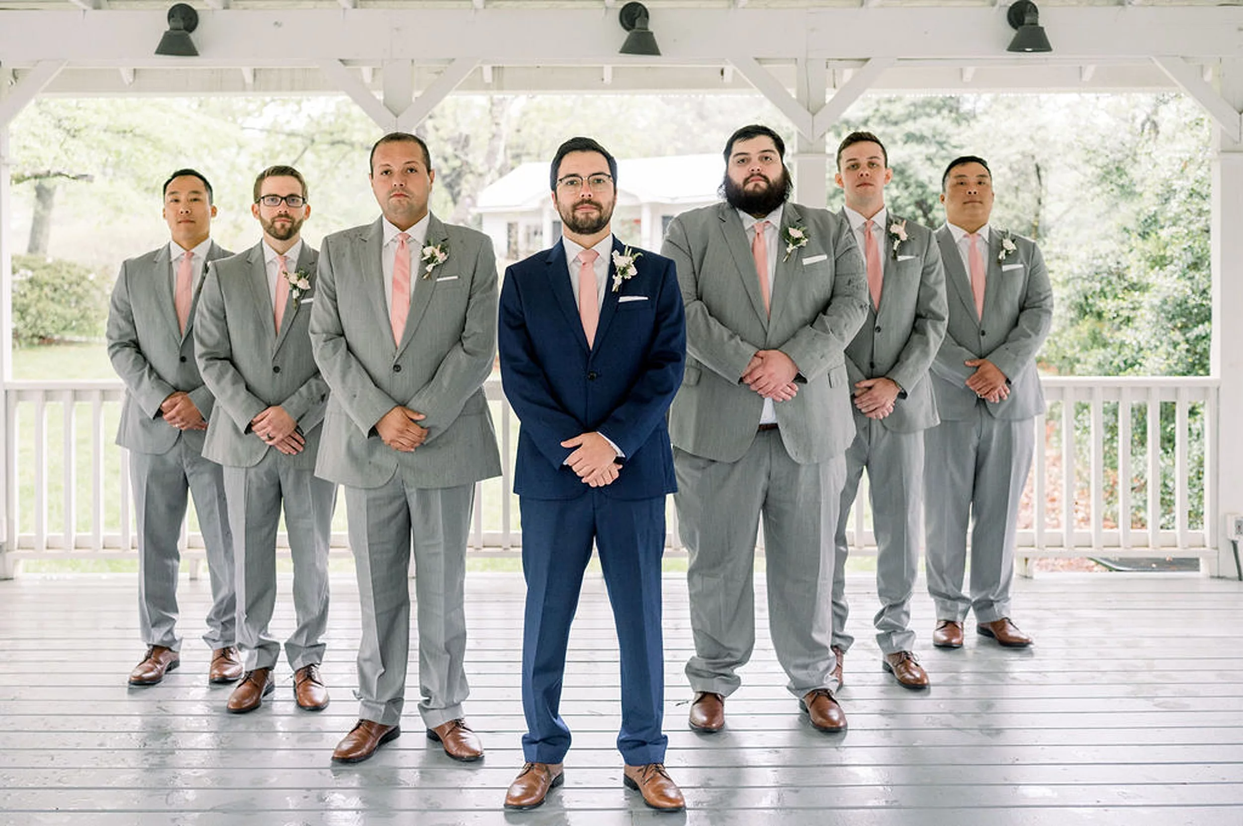 A groom in a blue suit stands with hands folded in front of him with his six groomsman in grey suits in a "V" behind him standing on a porch at a payne-corley house wedding