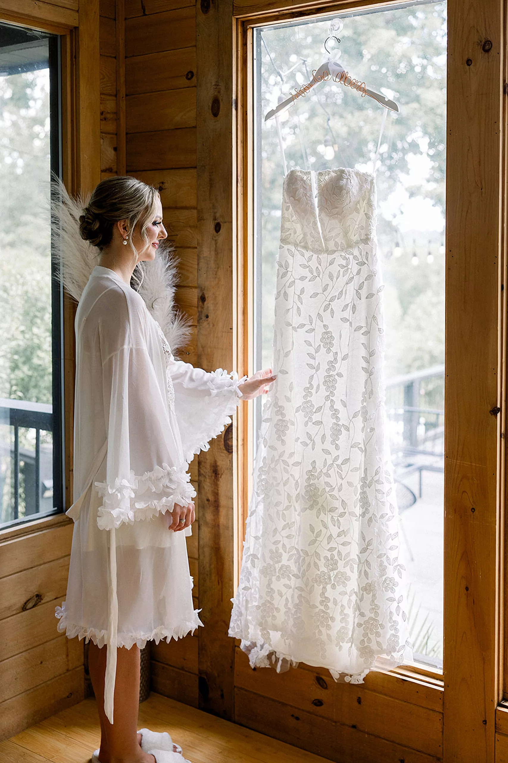 A bride in white pajamas looks at her dress as it hangs in a window from a custom hanger