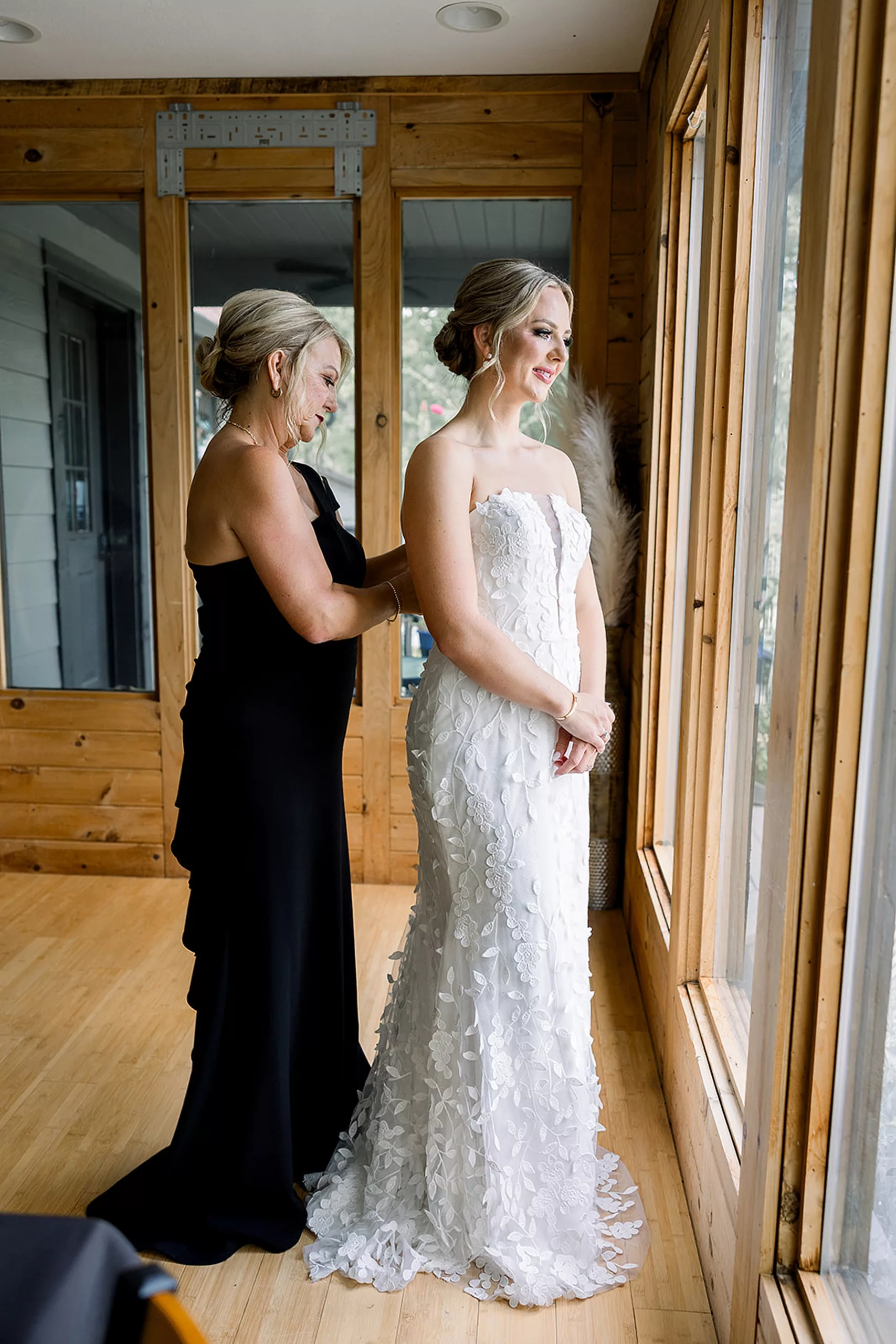 A bride stands in a window as her mother in a black dress buttons up her dress