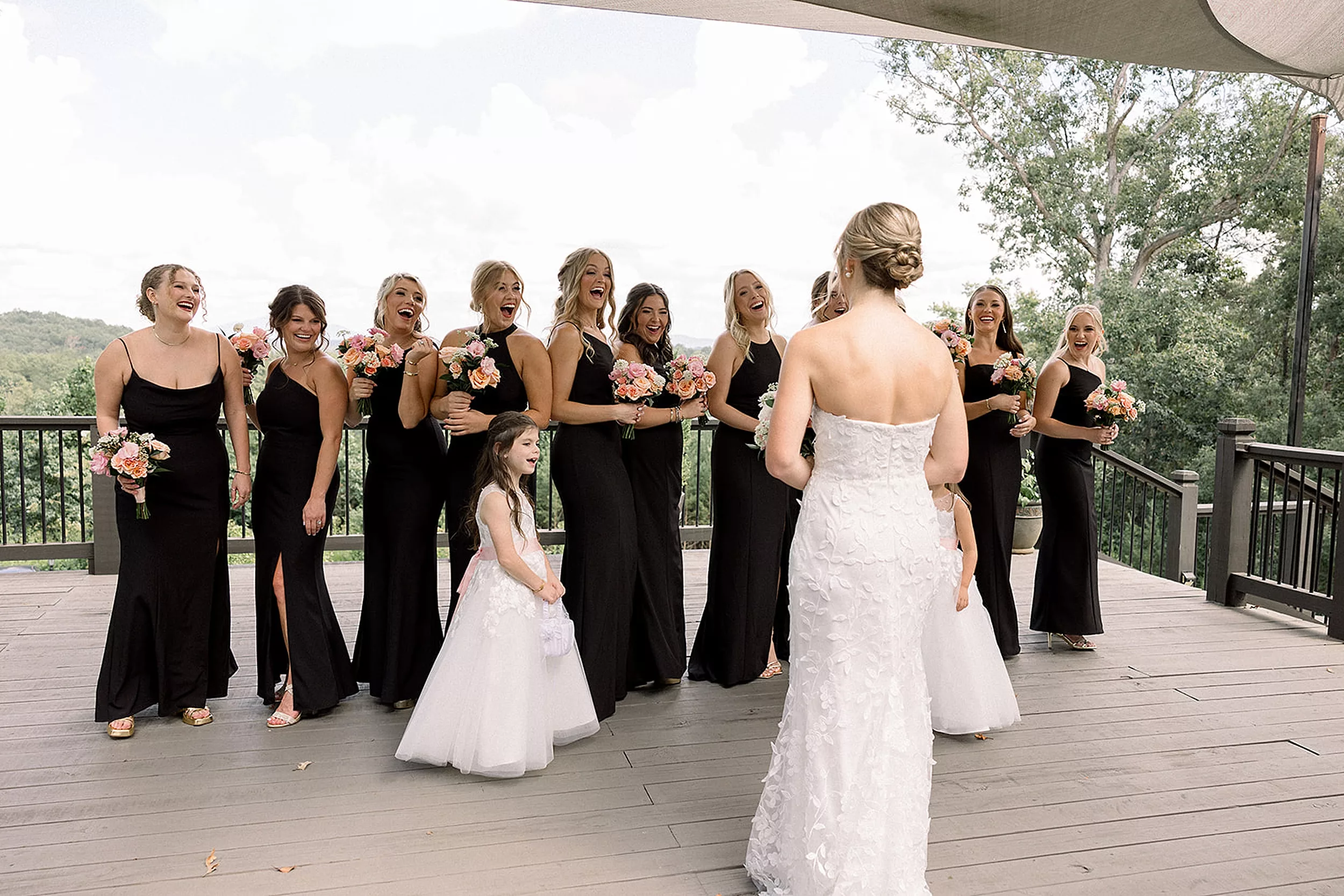 A bridal party in all black dresses stands on a porch and celebrates as they see the bride in her dress for the first time